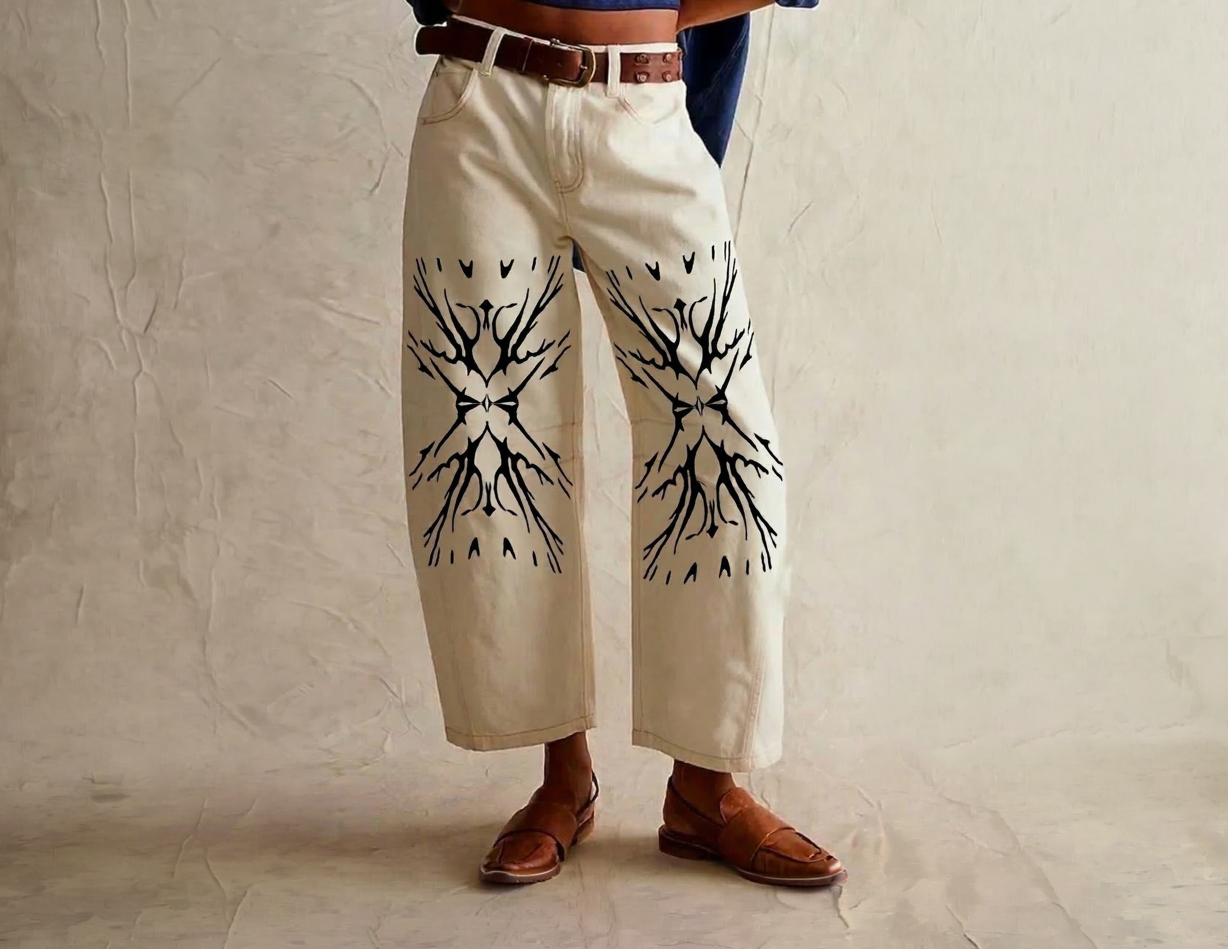 Person wearing beige pants with black pattern designs on the front, brown belt, brown shoes, and a dark blue jacket against a plain wall background.