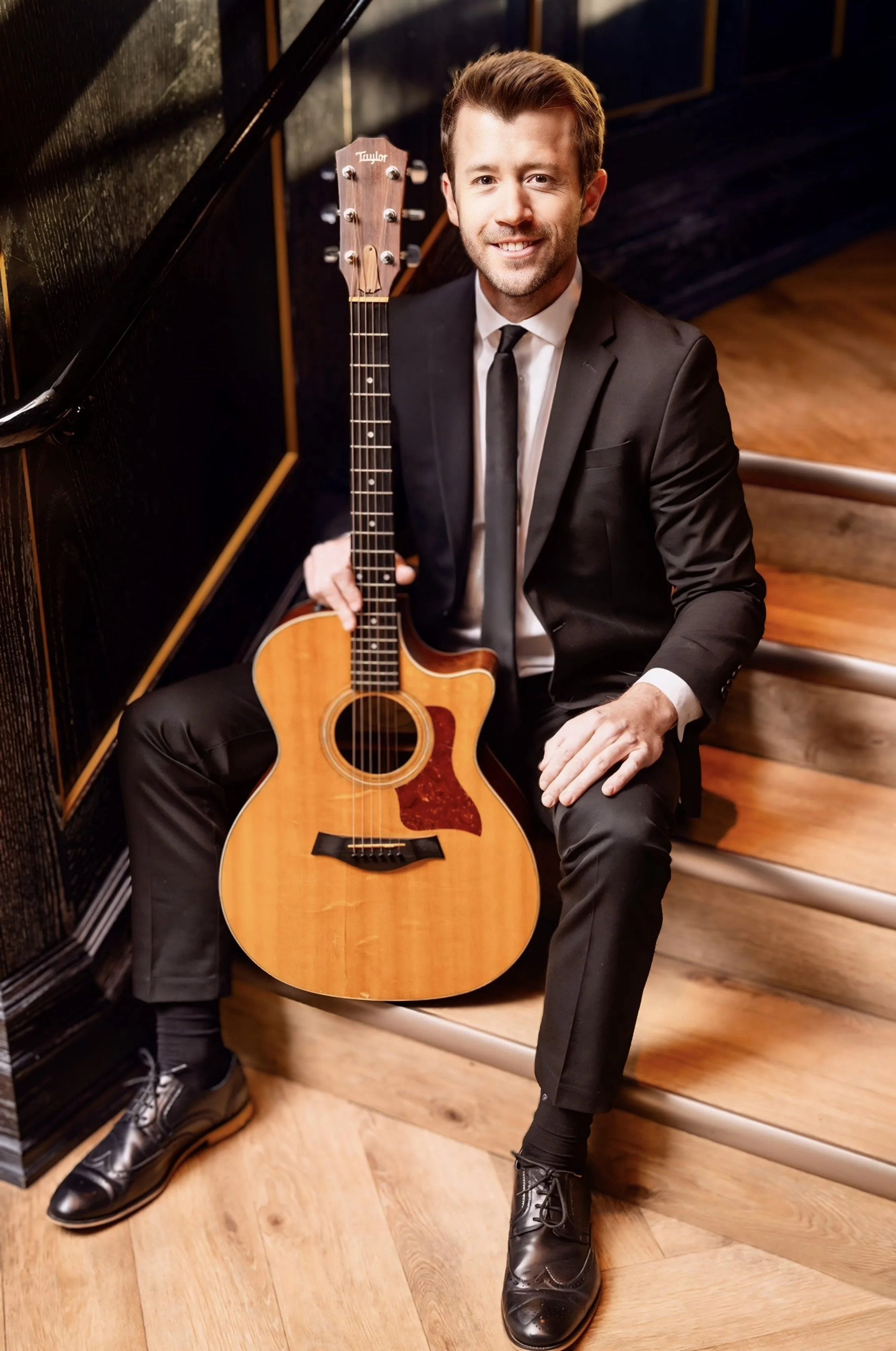 A man in a black suit and tie sitting on wooden stairs, holding an acoustic guitar, smiling at the camera.