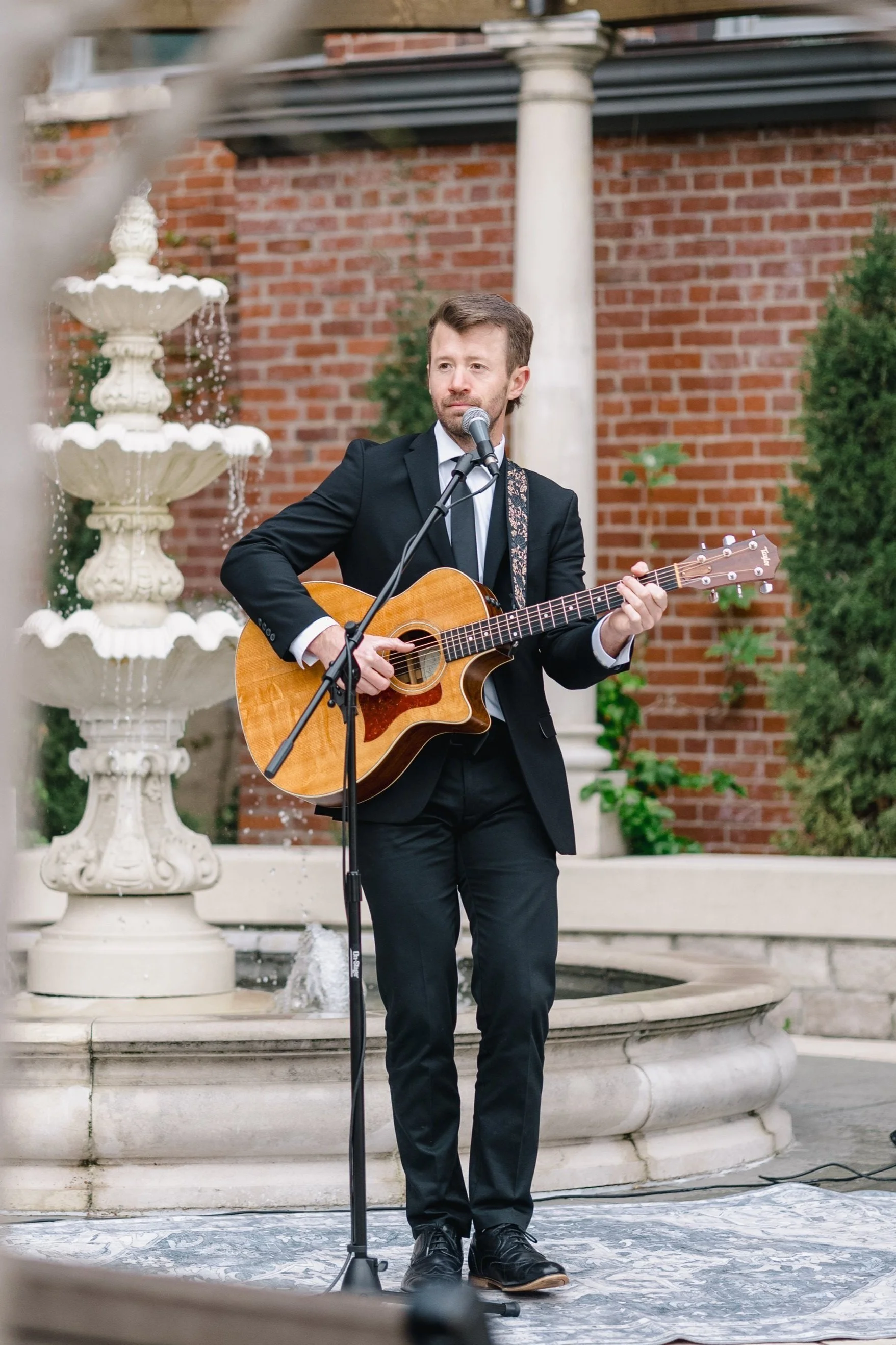 A man in a black suit playing an acoustic guitar and singing into a microphone in an outdoor setting near a fountain and brick building.