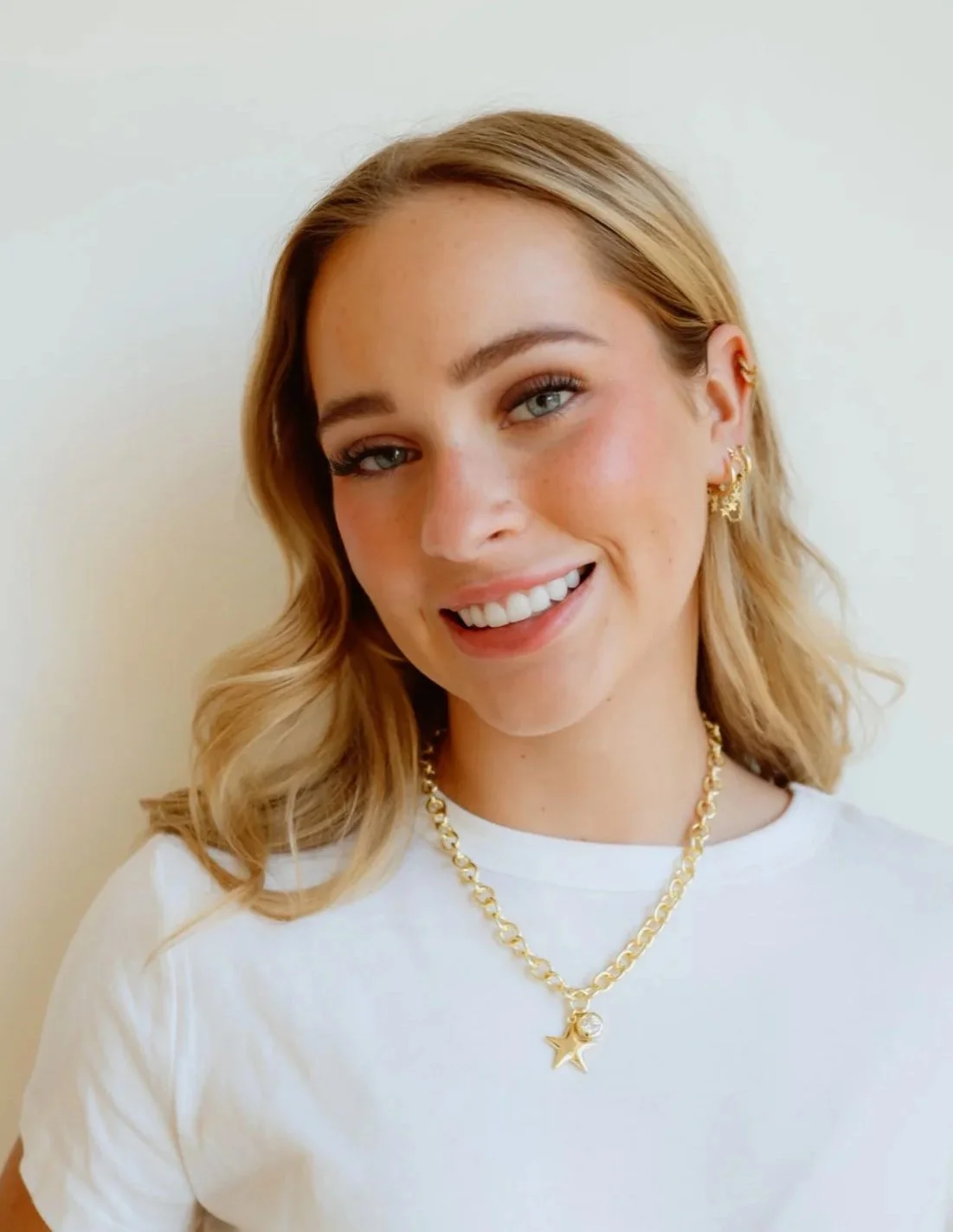 A young woman with blonde hair, blue eyes, and light skin, smiling at the camera, wearing a white t-shirt, gold star-shaped jewelry, and a gold chain necklace with a star pendant, standing against a plain light-colored background.