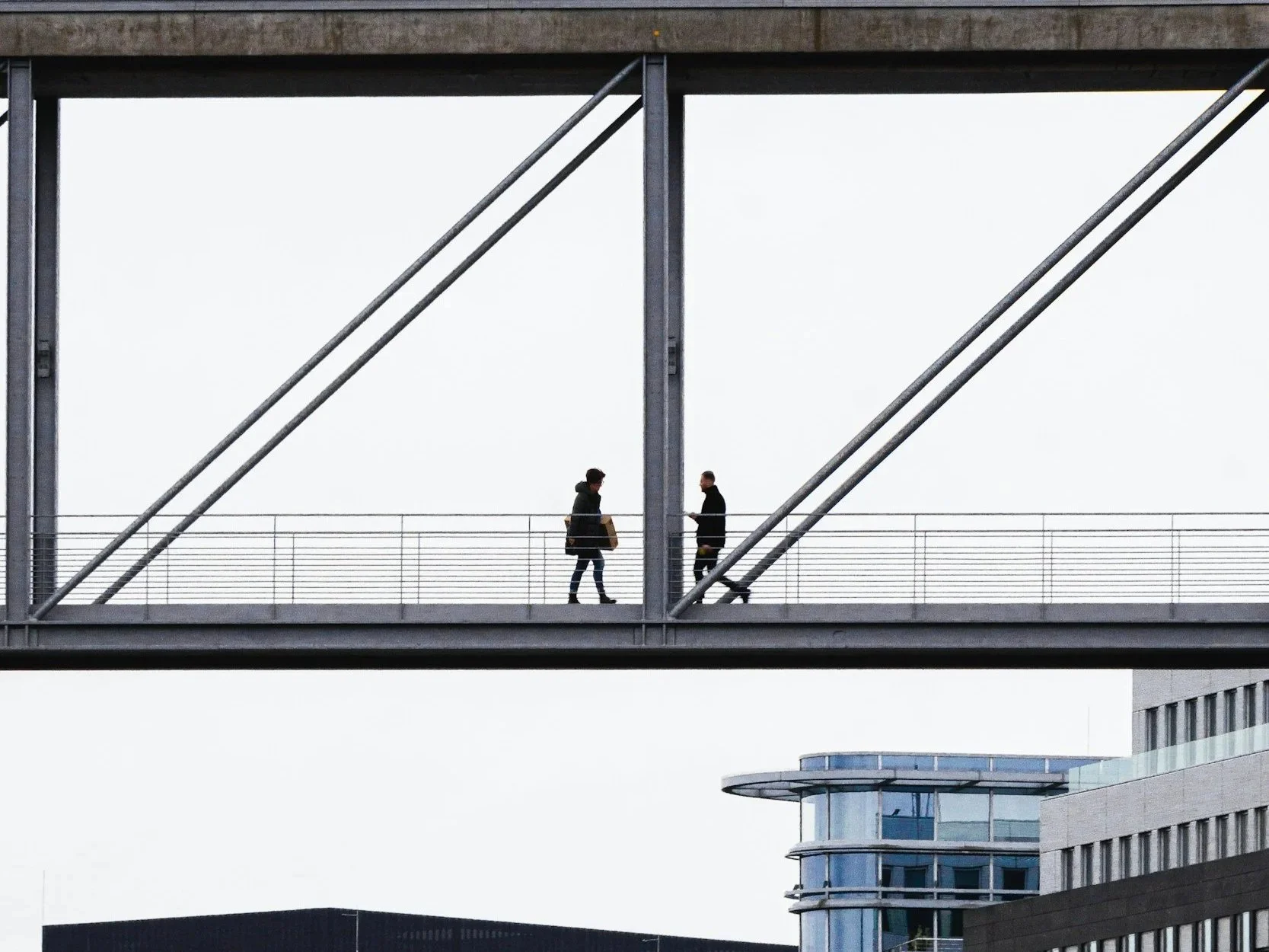 Two people walking on a modern, elevated pedestrian bridge with a metal railing, framed by a steel structure, in an urban setting.