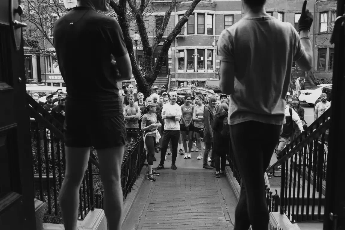 Two people standing on steps, addressing runners in an outdoor urban setting, Tracksmith's Boston Trackhouse, with buildings and trees in the background.