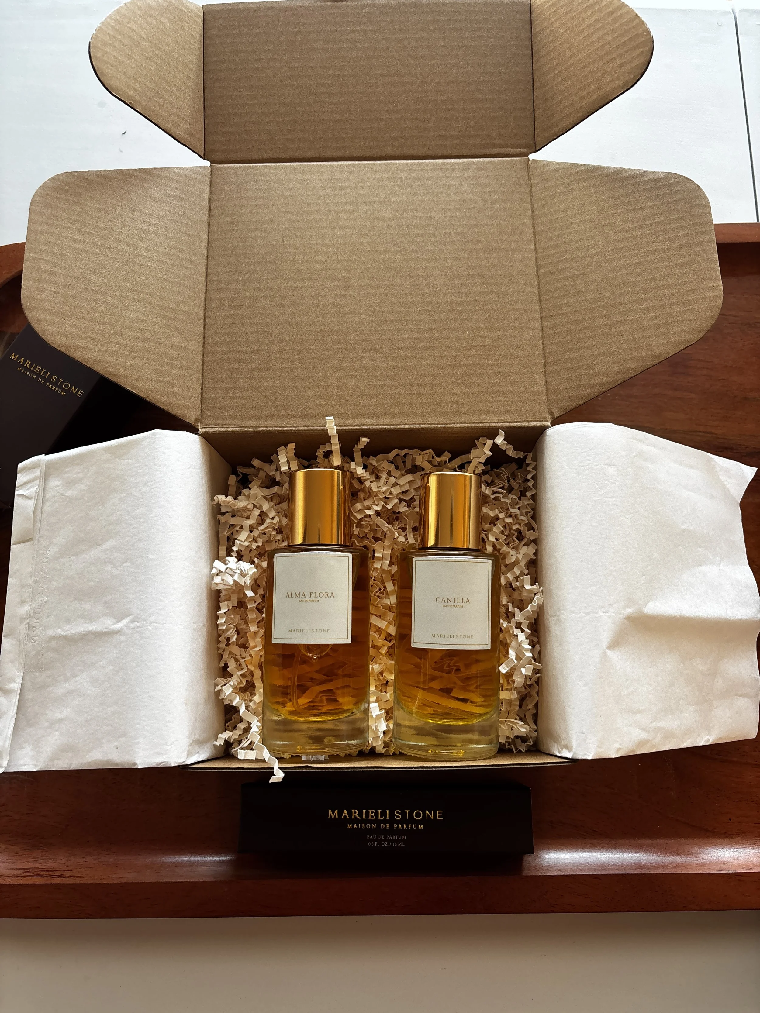 Open cardboard box containing two bottles of perfume labeled 'Alma Flora' and 'Canilla' with gold caps, surrounded by packing paper.