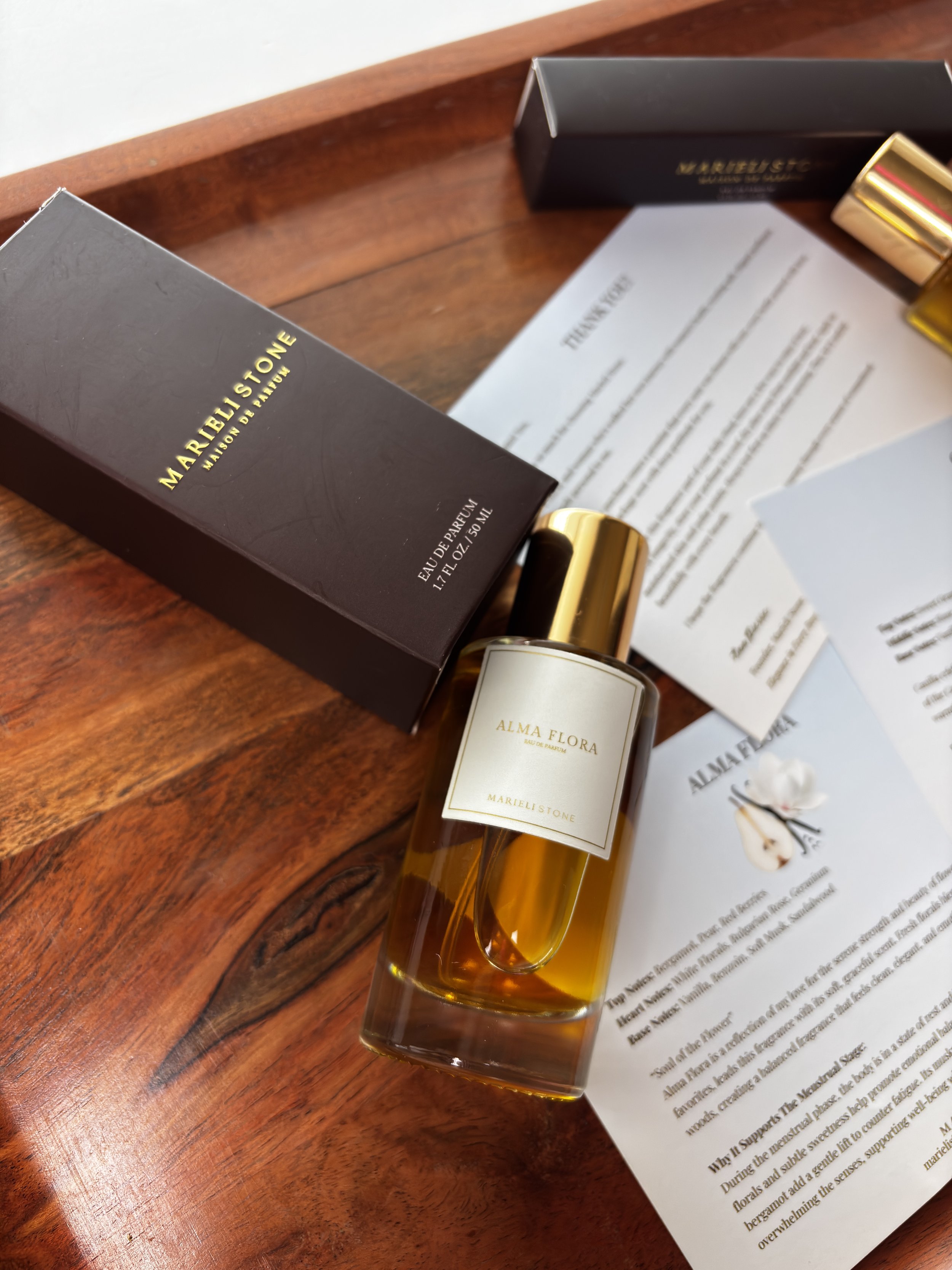 A bottle of Alma Flora perfume by Mariella Stone with its packaging and a product description sheet on a wooden surface.