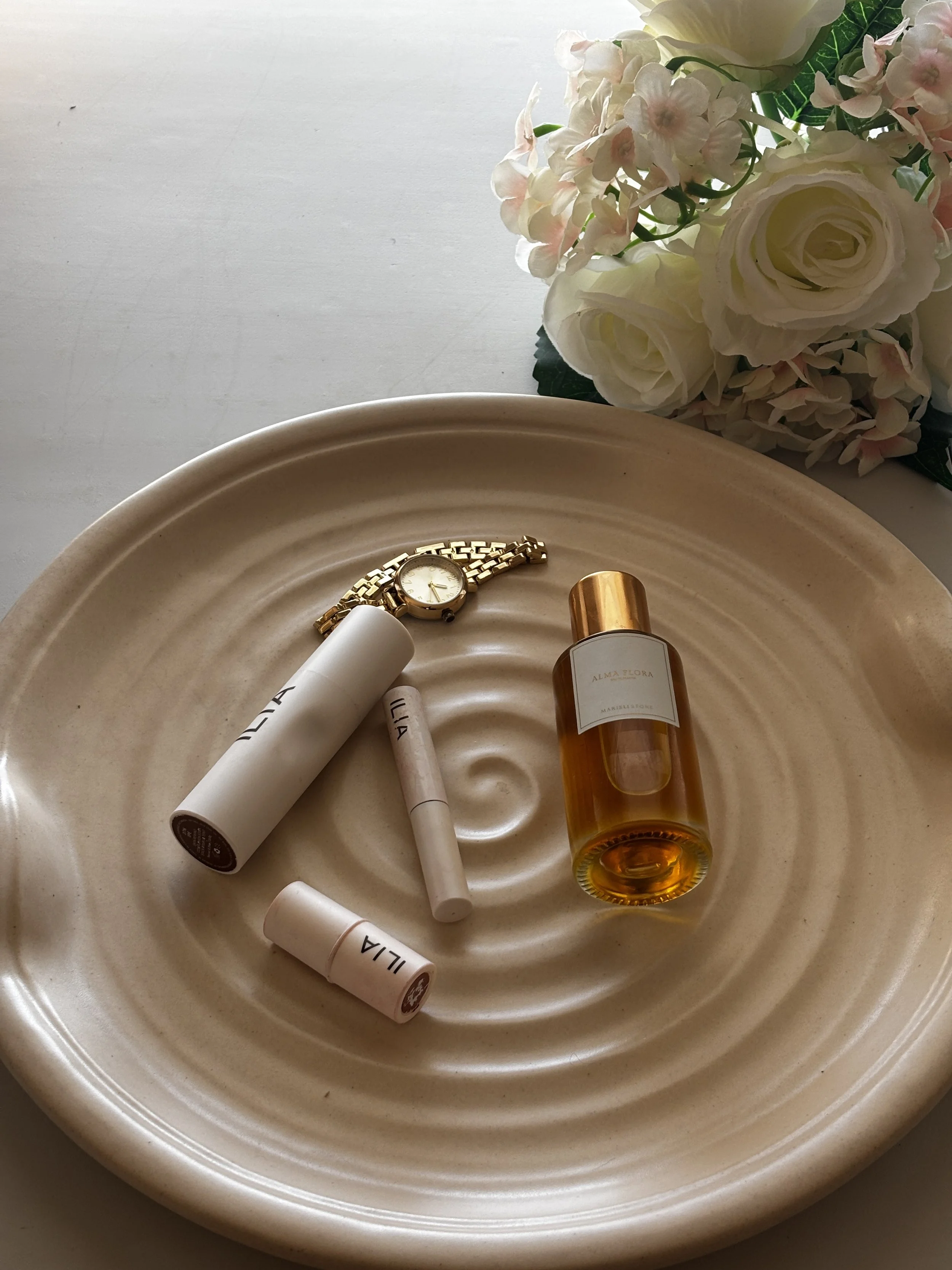 A beige ceramic plate holding a gold wristwatch, a perfume bottle, and several white skincare or cosmetic tubes. In the background, there is a bouquet of white and pink flowers.