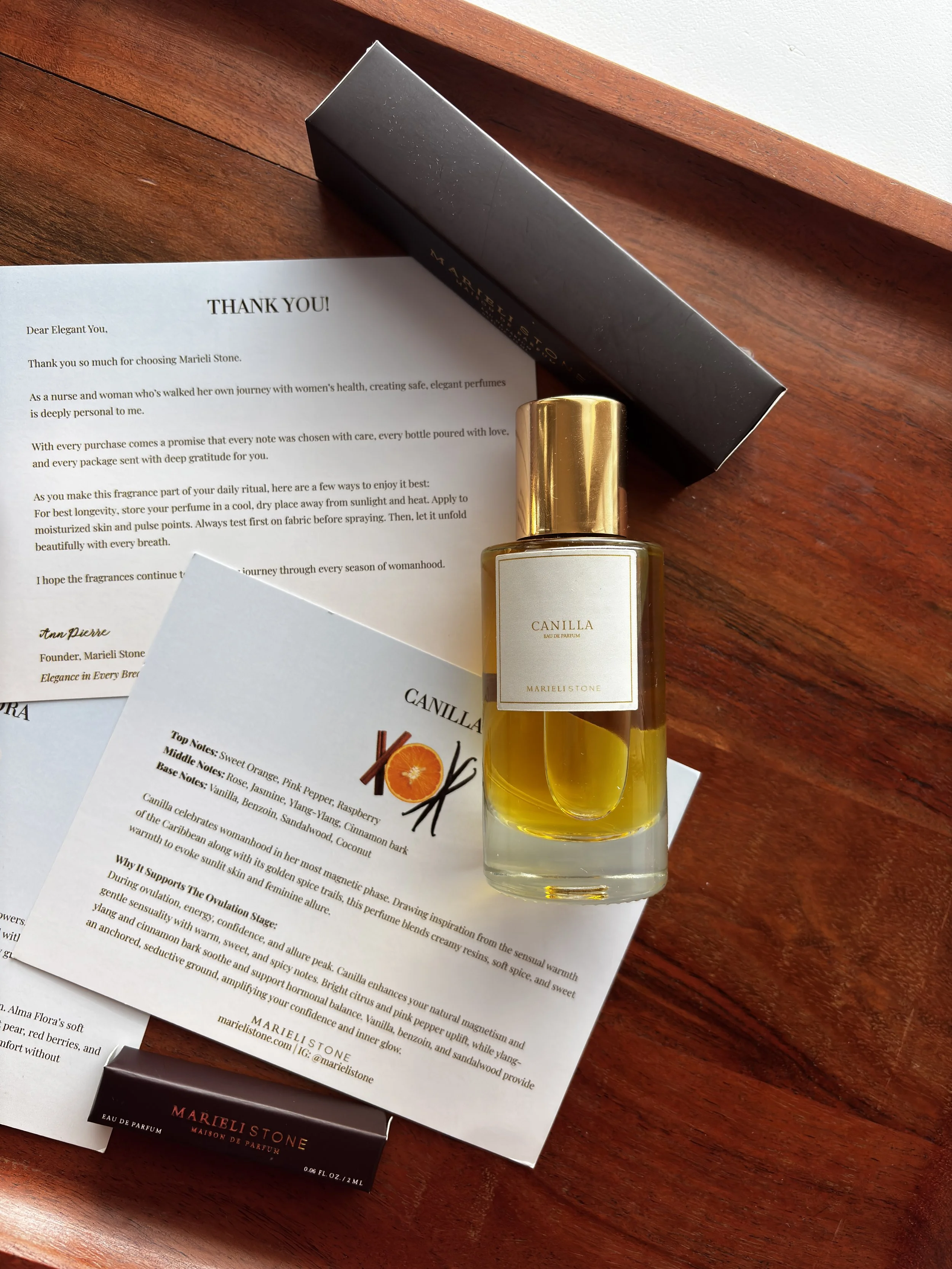 A bottle of Marieli Stone Canilla perfume, a black box, a small Marieli Stone perfume sample, and printed informational and thank you letters on a wooden surface.