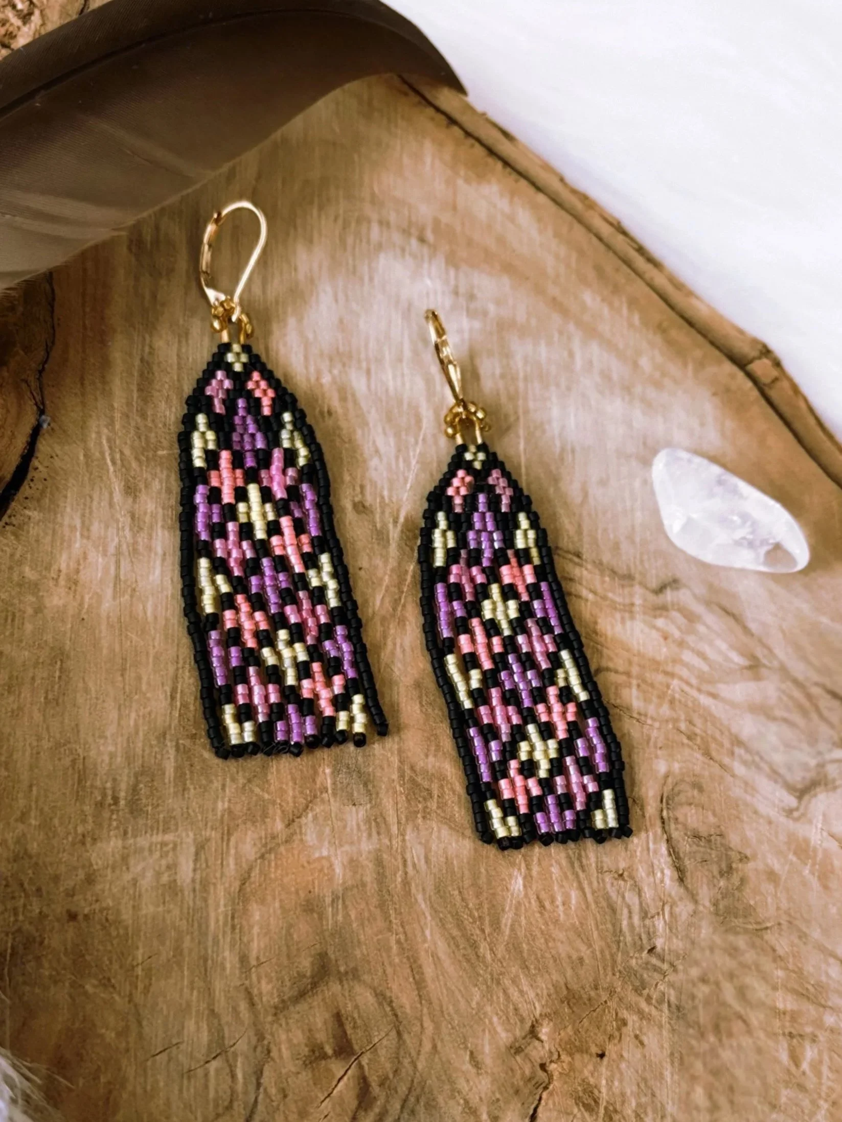 Beaded earrings with purple, pink, and black patterns resting on a wooden surface with white feathers and a small crystal nearby.