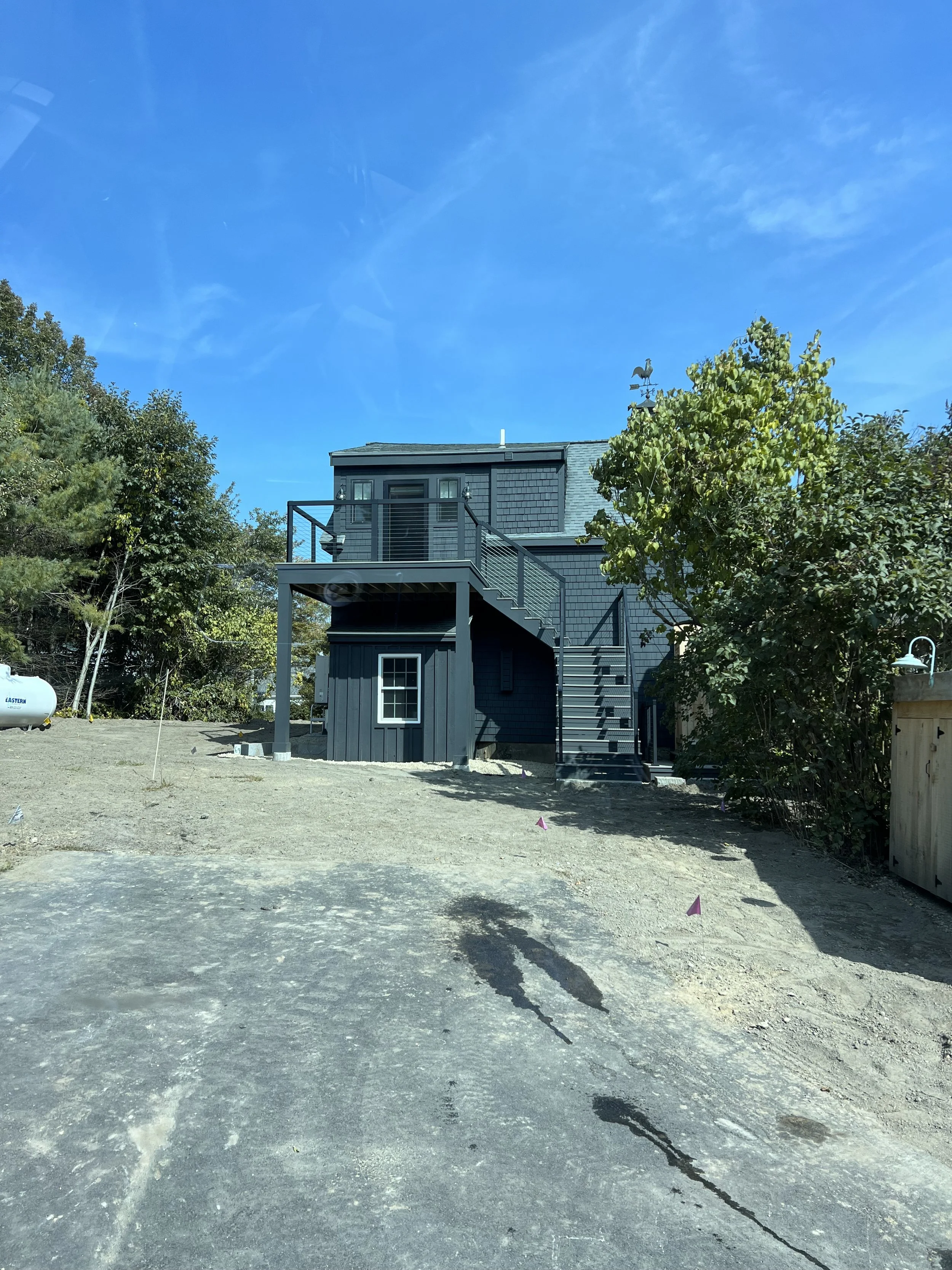 A modern two-story house painted in dark blue with an outdoor staircase leading to a second-floor balcony. The house has a small window on the ground level and a large window on the upper level. There are trees on both sides, a propane tank on the left, and a clear blue sky above.