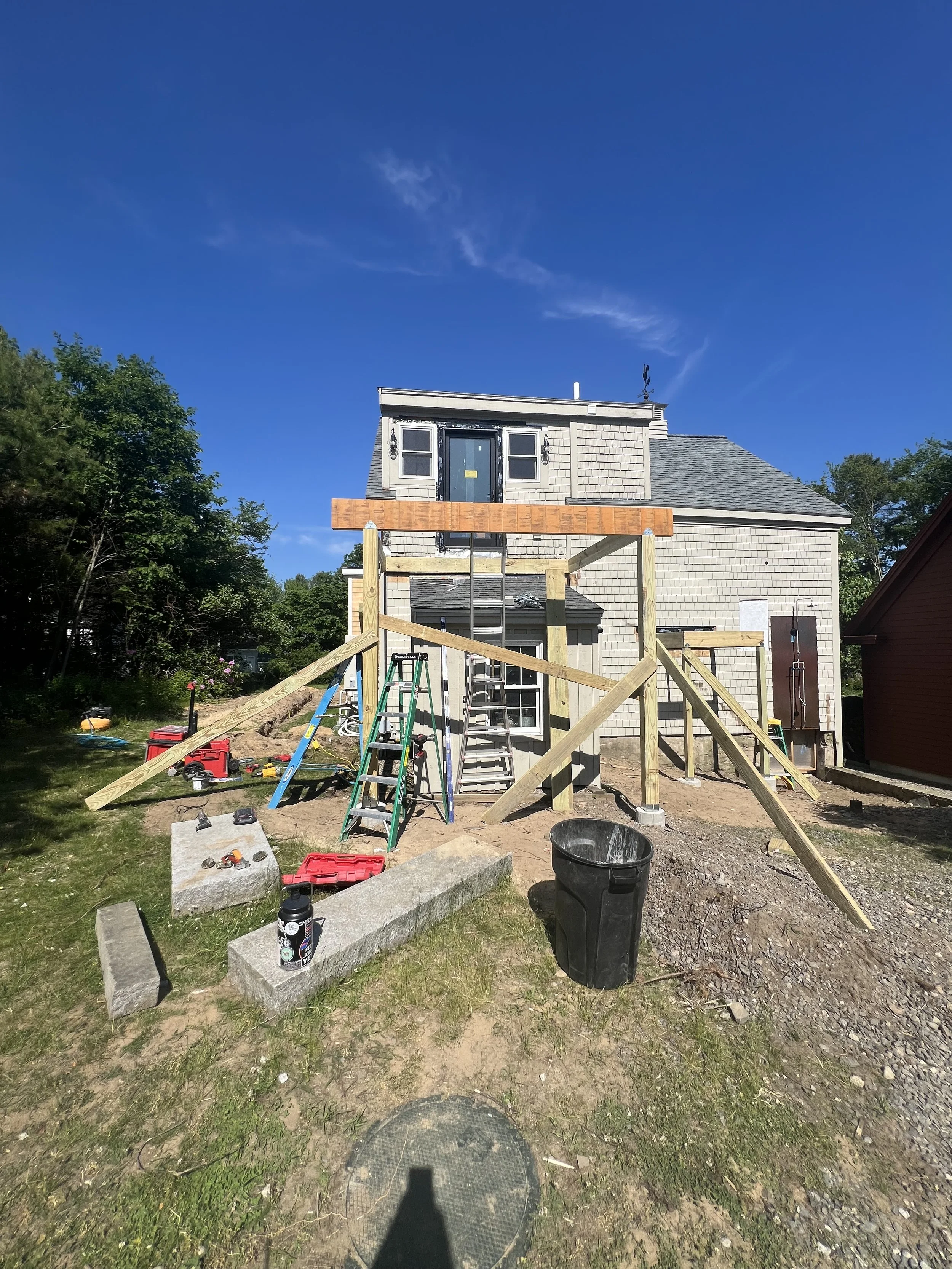 Backyard construction site with a partially built second-story deck or extension on a house, scaffolding, ladders, tools, and construction materials on a sunny day.