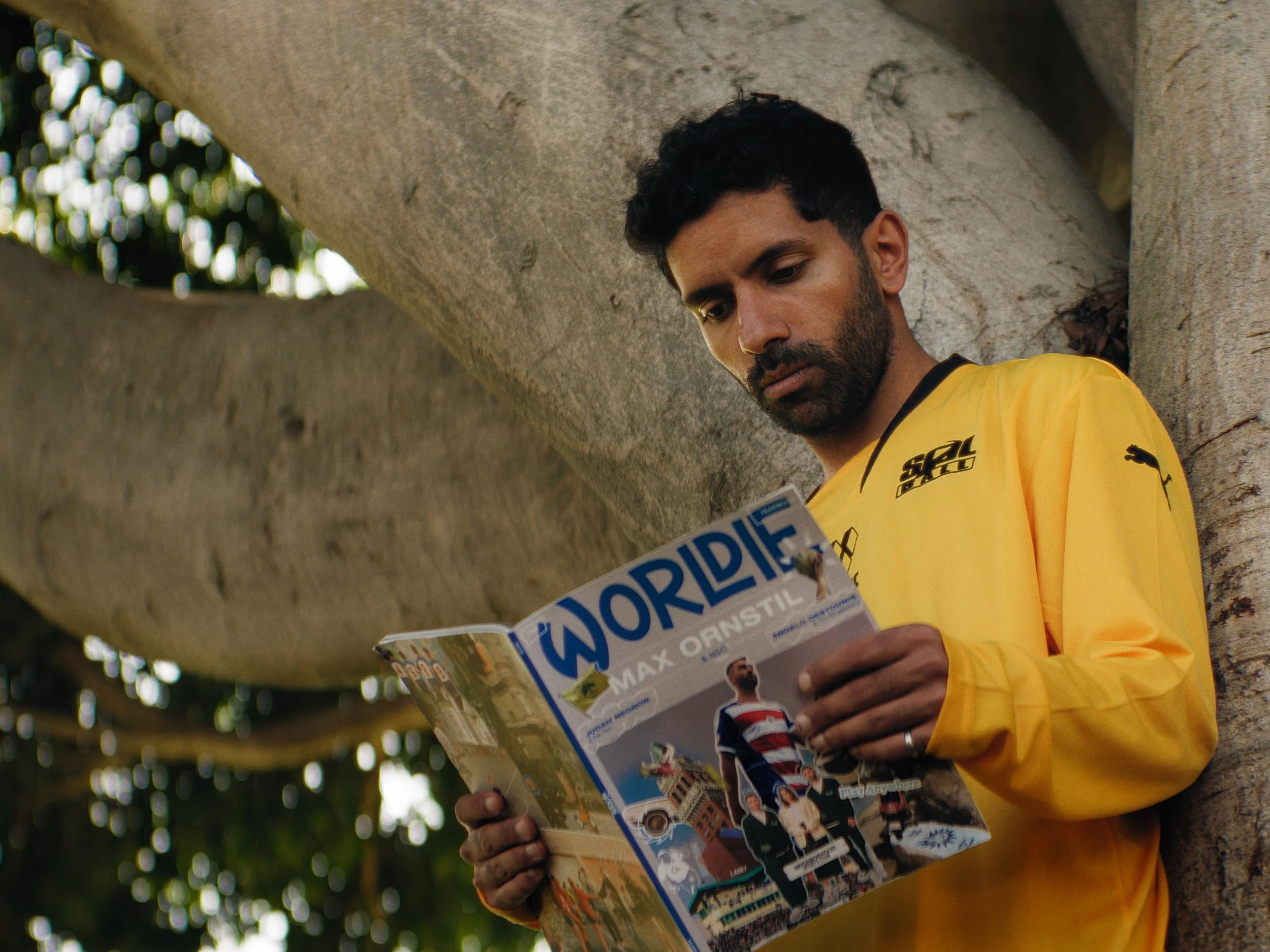 Man in yellow shirt leaning against tree while reading a magazine.