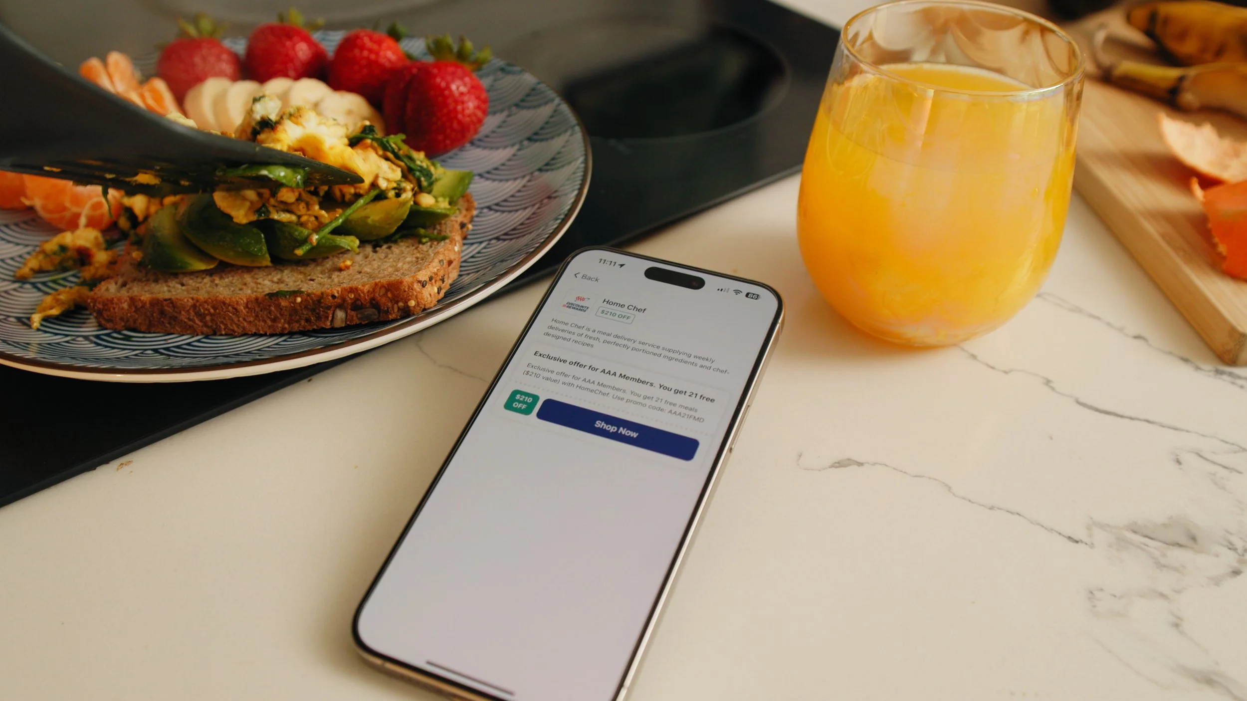 A breakfast scene with a plate of strawberries, scrambled eggs with spinach on whole grain toast, a glass of orange juice, a cutting board with bananas and sliced fruit, and a smartphone displaying a Home Chef promotion on a marble countertop.