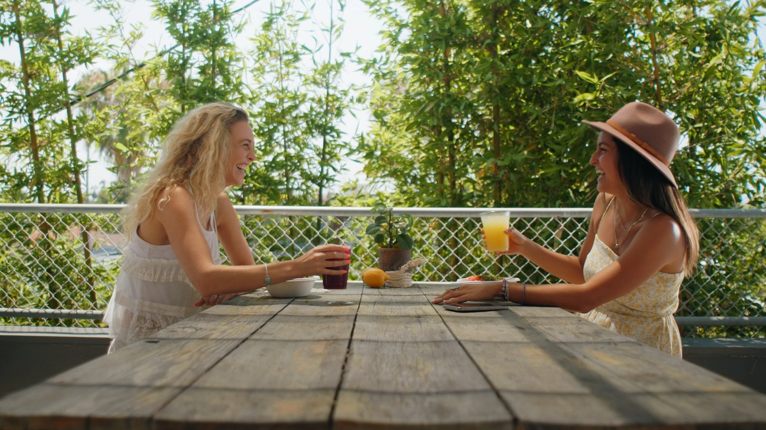 Two women sitting at a wooden table outdoors, smiling and talking. One woman with long blonde hair wearing a white dress, the other with long dark hair wearing a tan hat and a yellow dress, holding drinks. The table has a potted plant, lemons, and bo