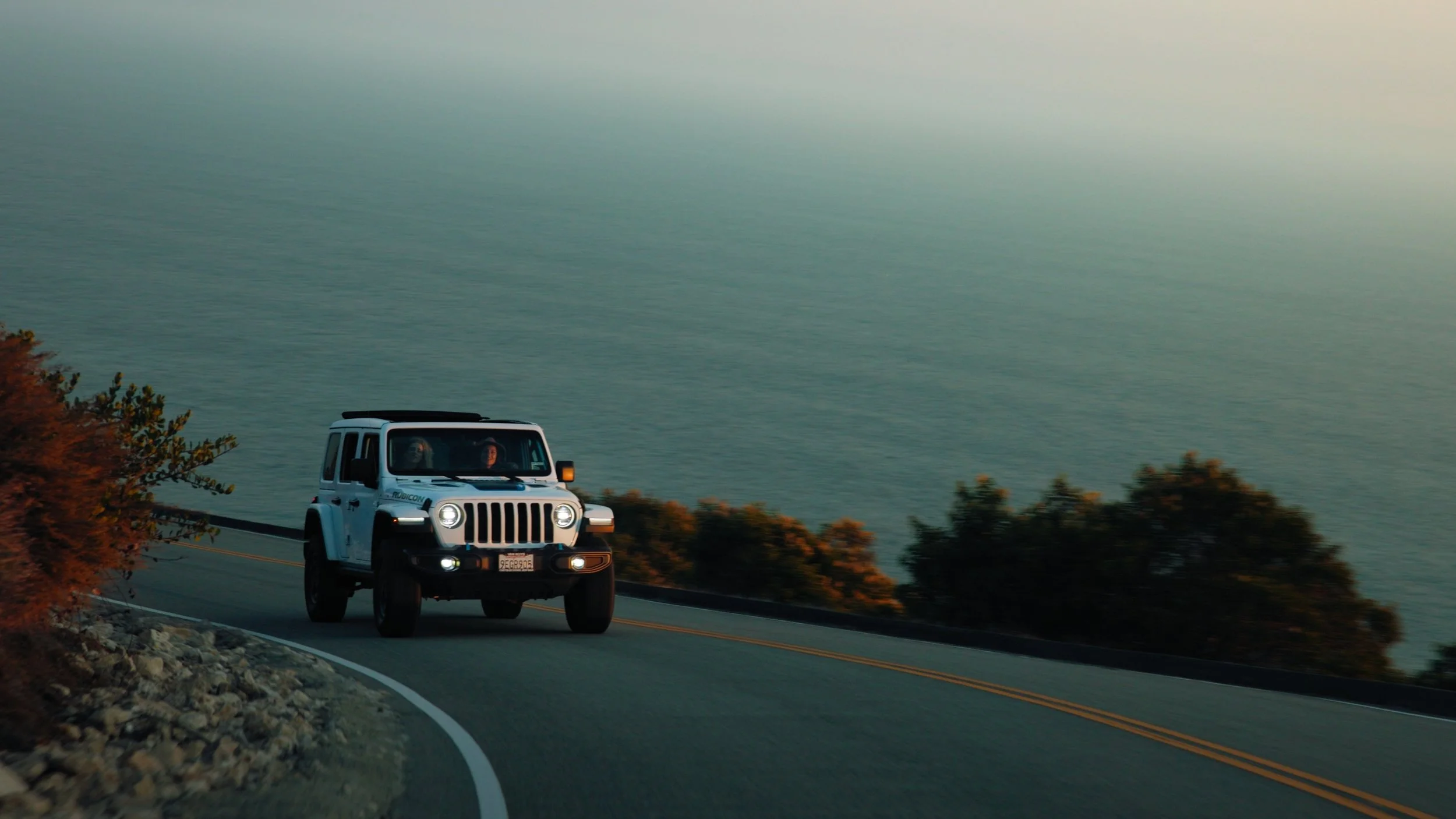 A white Jeep Wrangler driving along a winding mountain road overlooking a body of water, with trees on either side and a sunset or early evening sky.