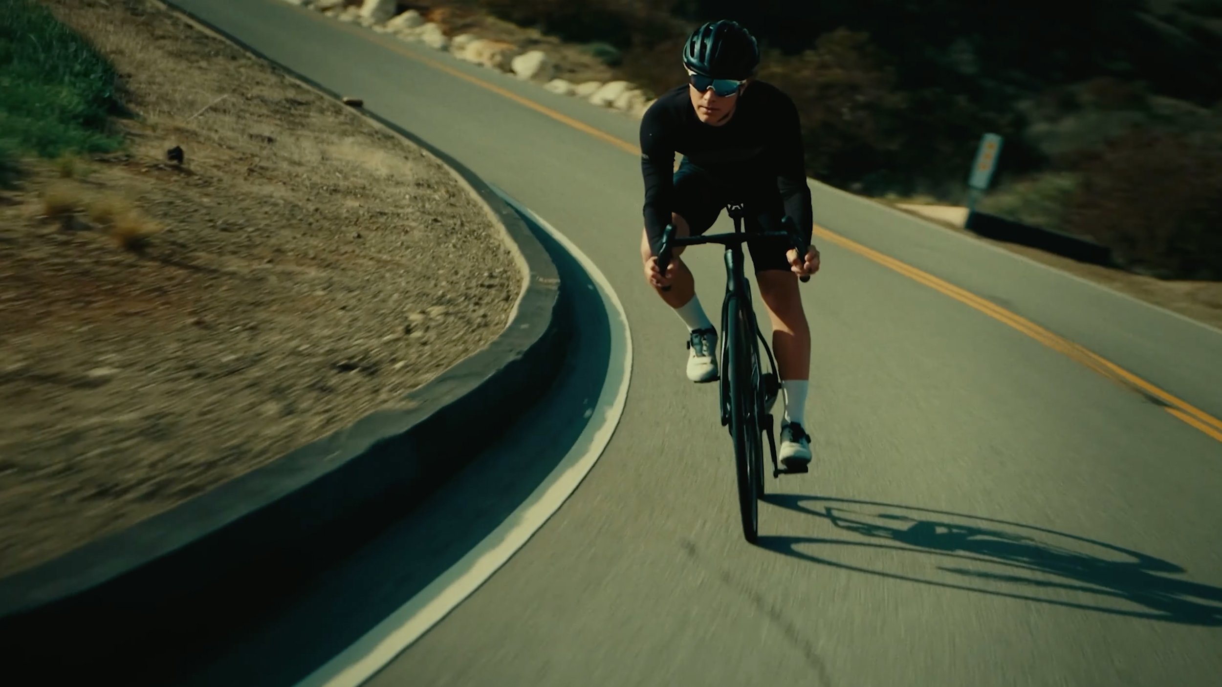 A man rides a black road bicycle on a winding mountain road, wearing a black helmet, sunglasses, black cycling jersey, and shorts.