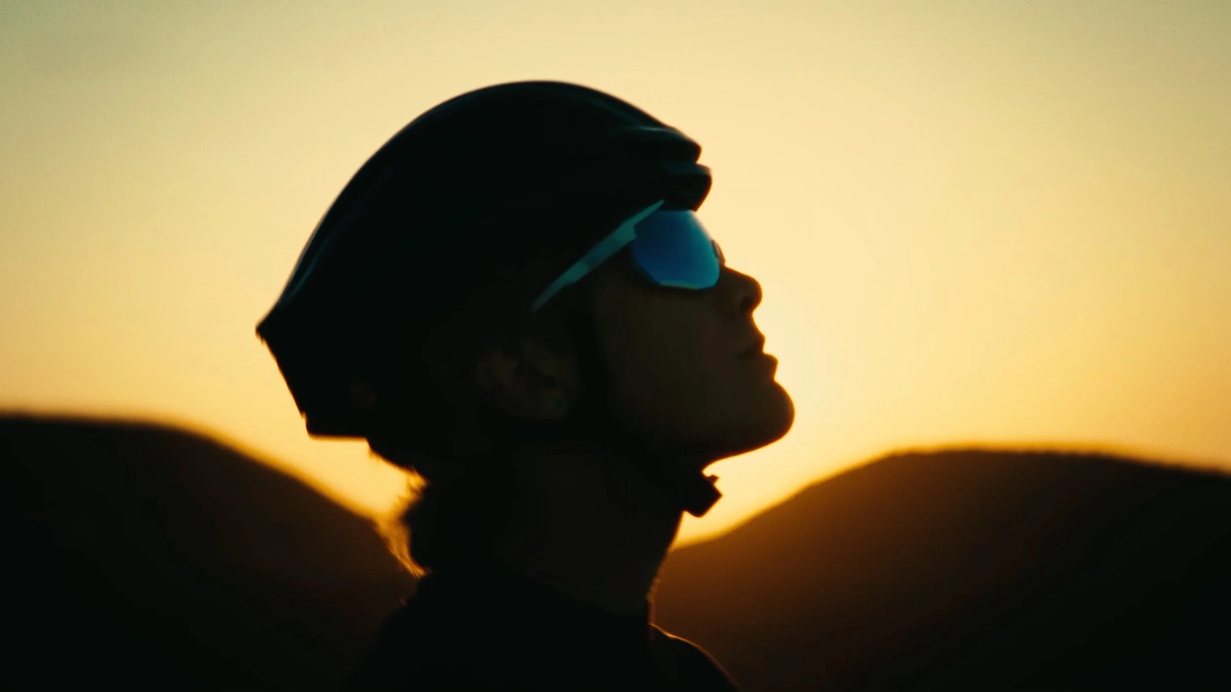 Silhouette of a person wearing a helmet and sunglasses, looking up against a sunset background with mountains.