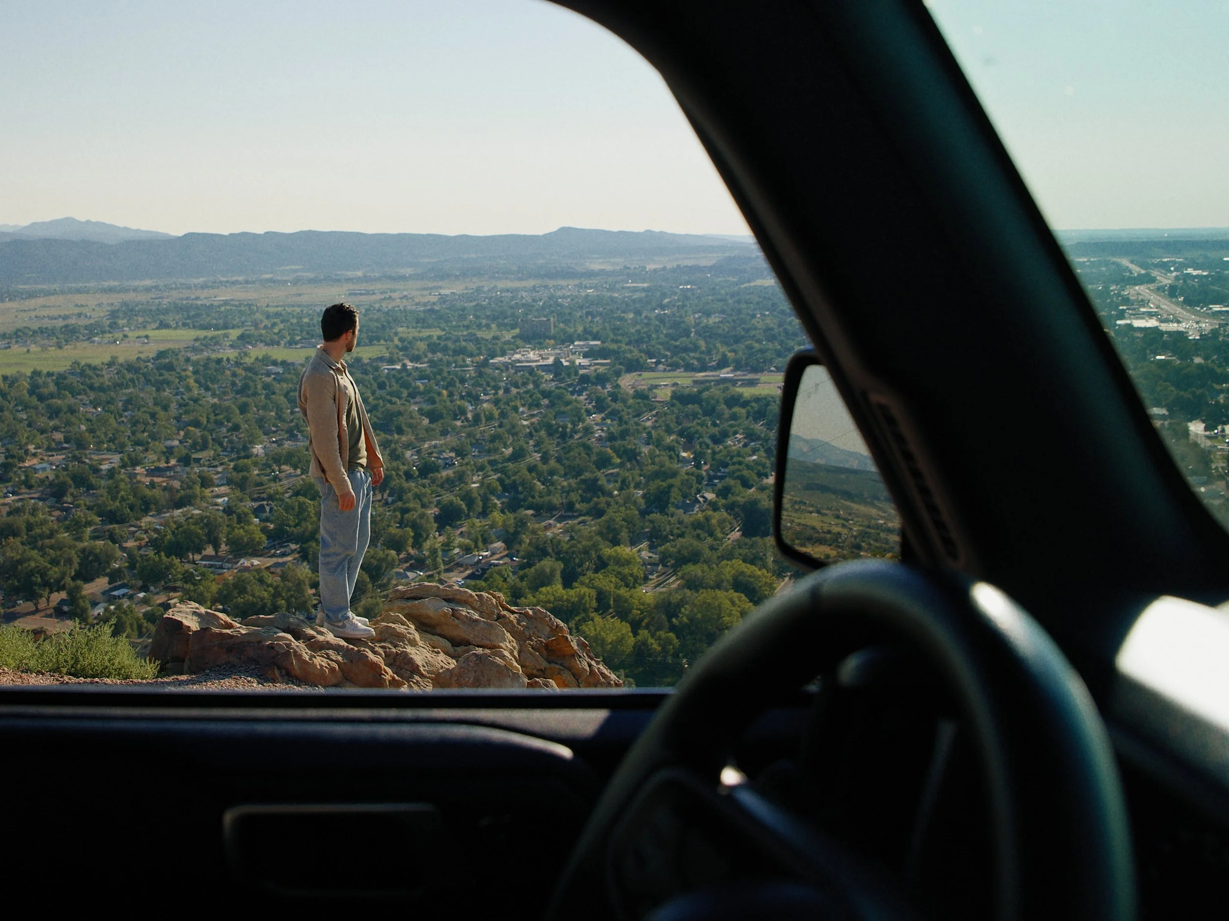 A person standing on a rocky ledge overlooking a city and landscape, viewed from inside a vehicle.
