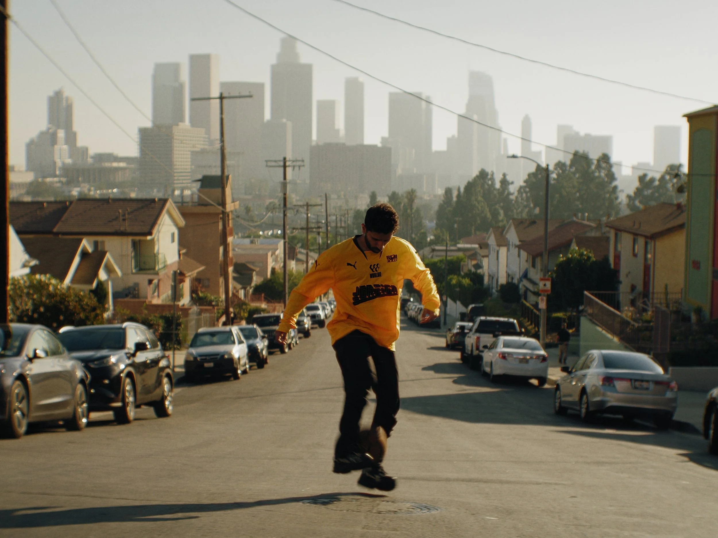A young man skating down a residential street with parked cars on both sides, houses on the hill, utility poles, and a city skyline in the background in Los Angeles.