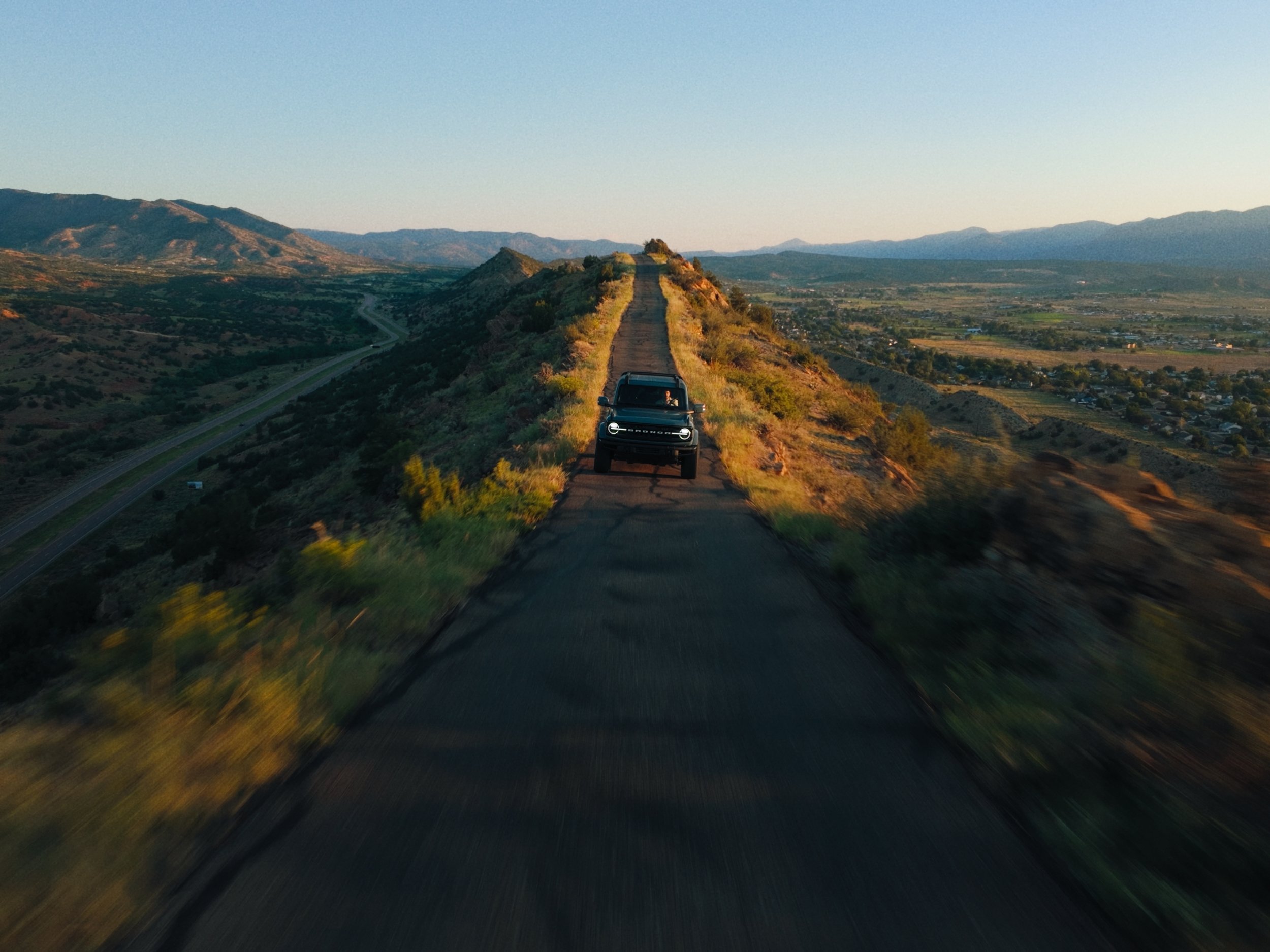 A black vehicle driving on a narrow, elevated road through mountainous terrain at sunset.