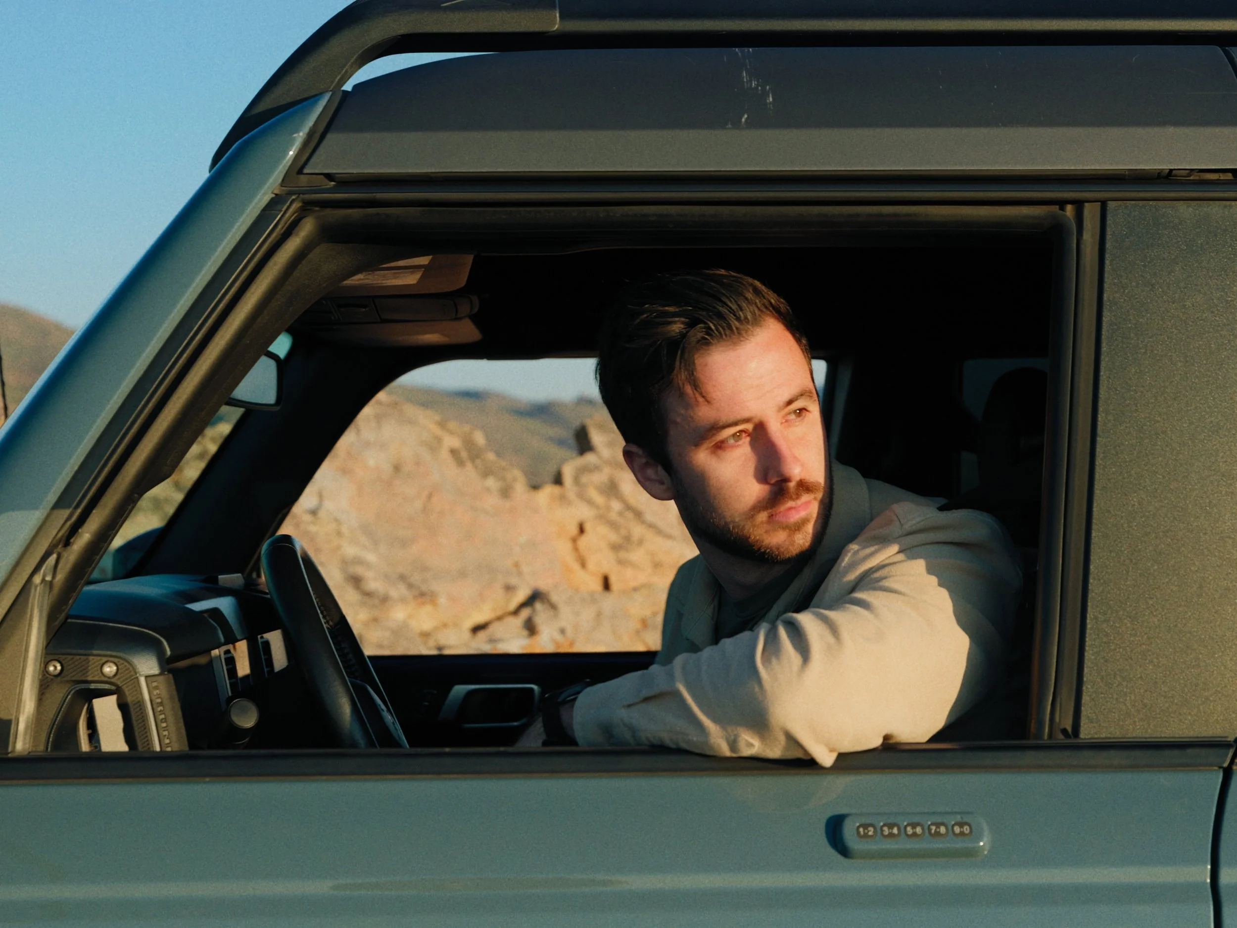 A man with dark hair and a beard sitting in the driver's seat of a green vehicle, looking out the window at a rocky desert landscape in the distance during sunset.