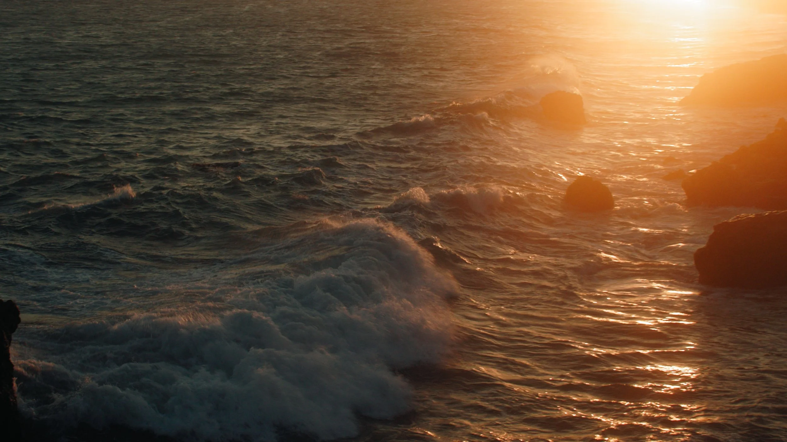 Ocean waves crashing against rocks during a sunset with sunlight reflecting on the water.