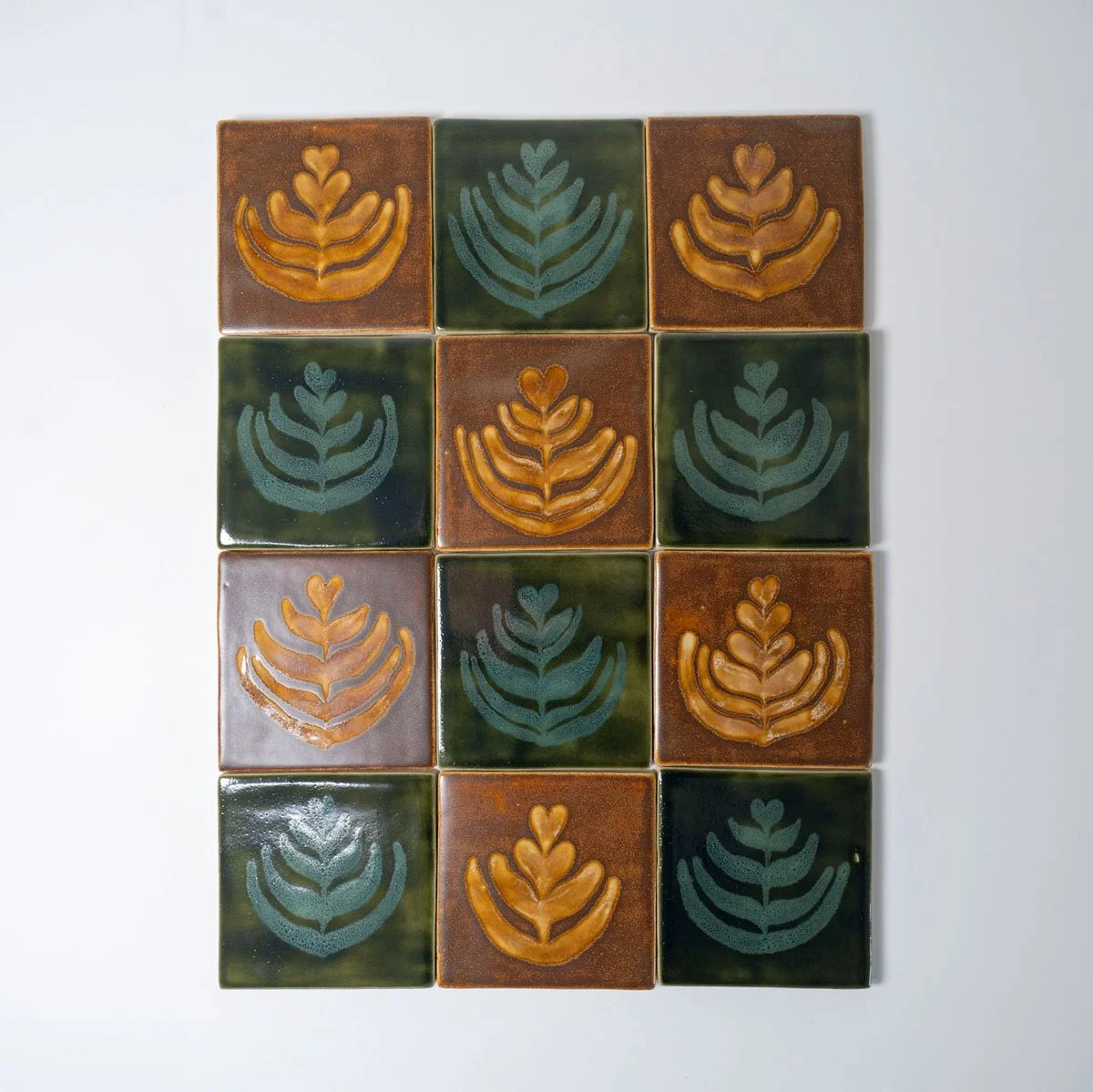 A square ceramic tile with a brown glaze and a latte art design resembling a leaf or fern in cream-colored foam.