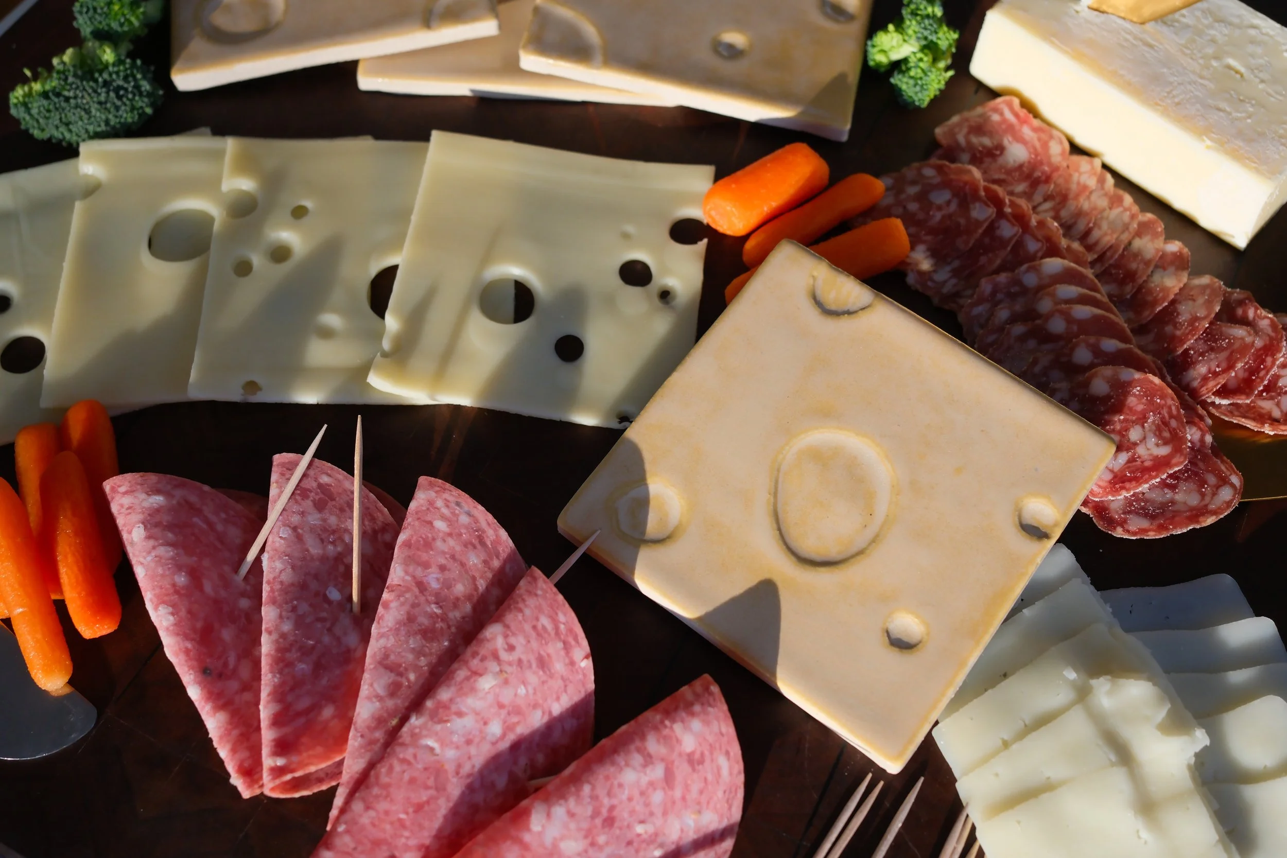 A photo of a swiss cheese themed ceramic tile on a charcuterie board surrounded by cheese and meat 