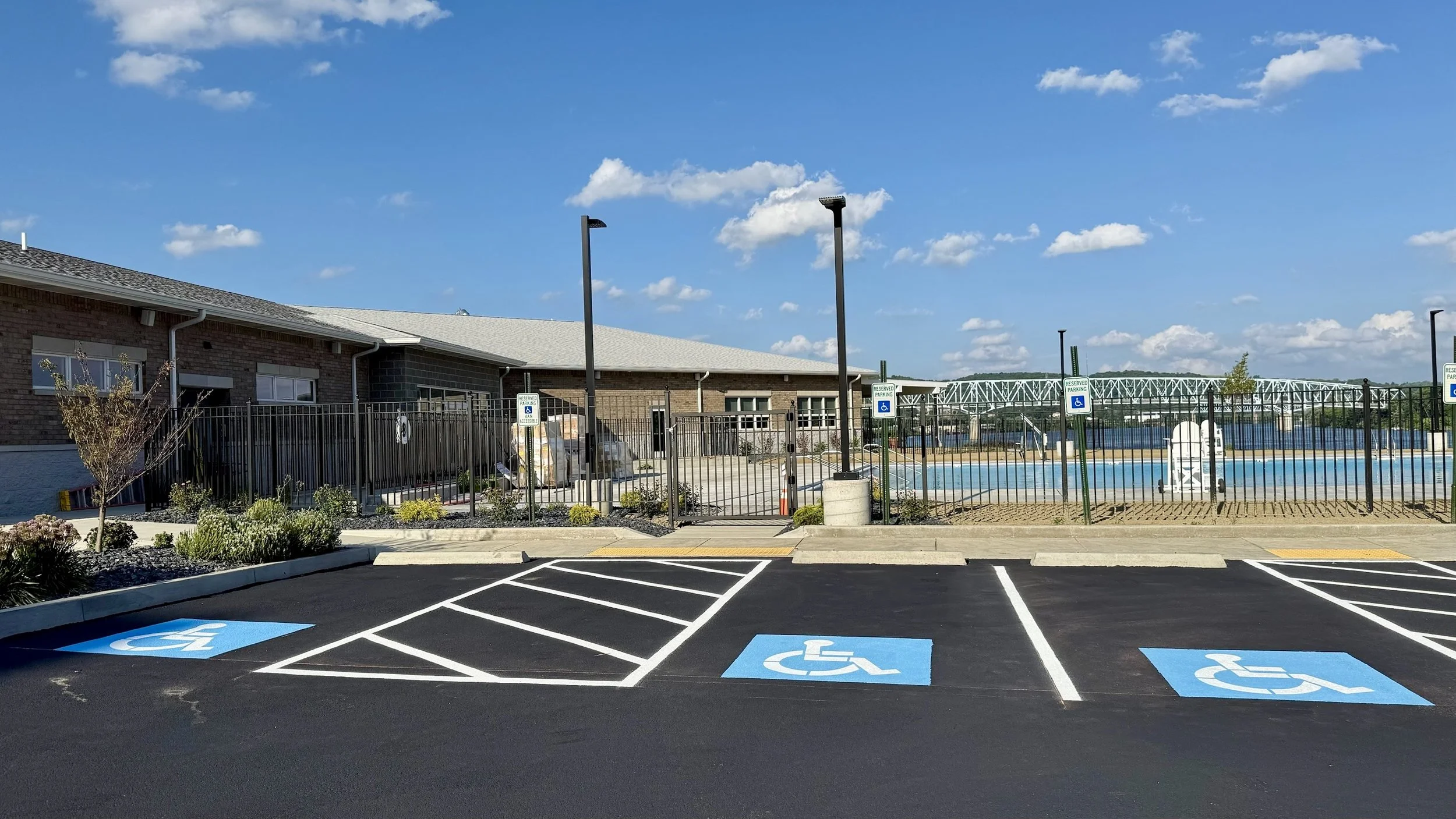 Parking lot with designated handicapped spaces in front of a pool area, which is enclosed by a fence and accessible through a gate, under a blue sky with scattered clouds.