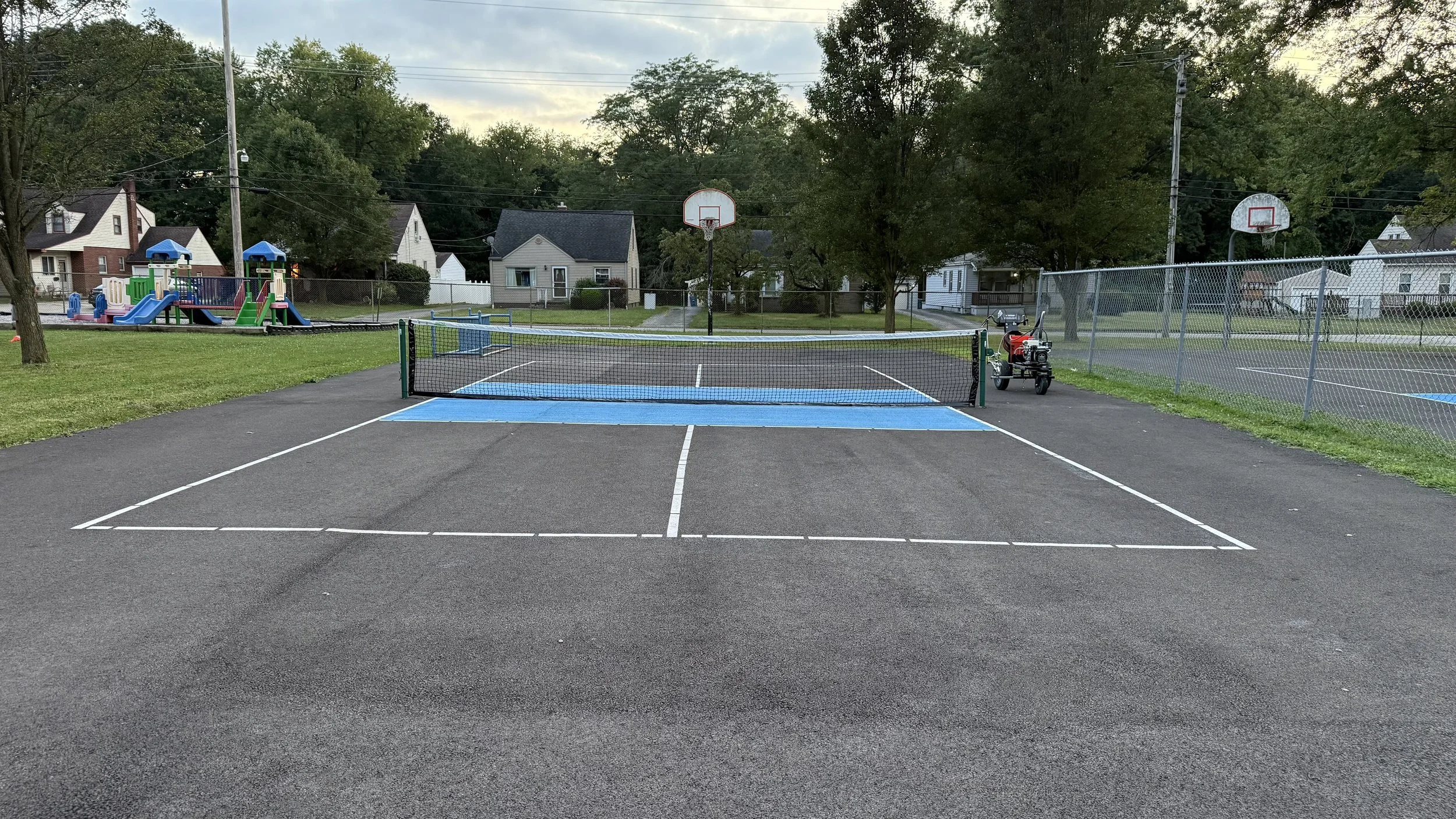 Empty park tennis court with a portable electric vehicle parked on the right, a playground with slides and climbing structures on the left, and two basketball hoops in the background, surrounded by trees and residential houses.