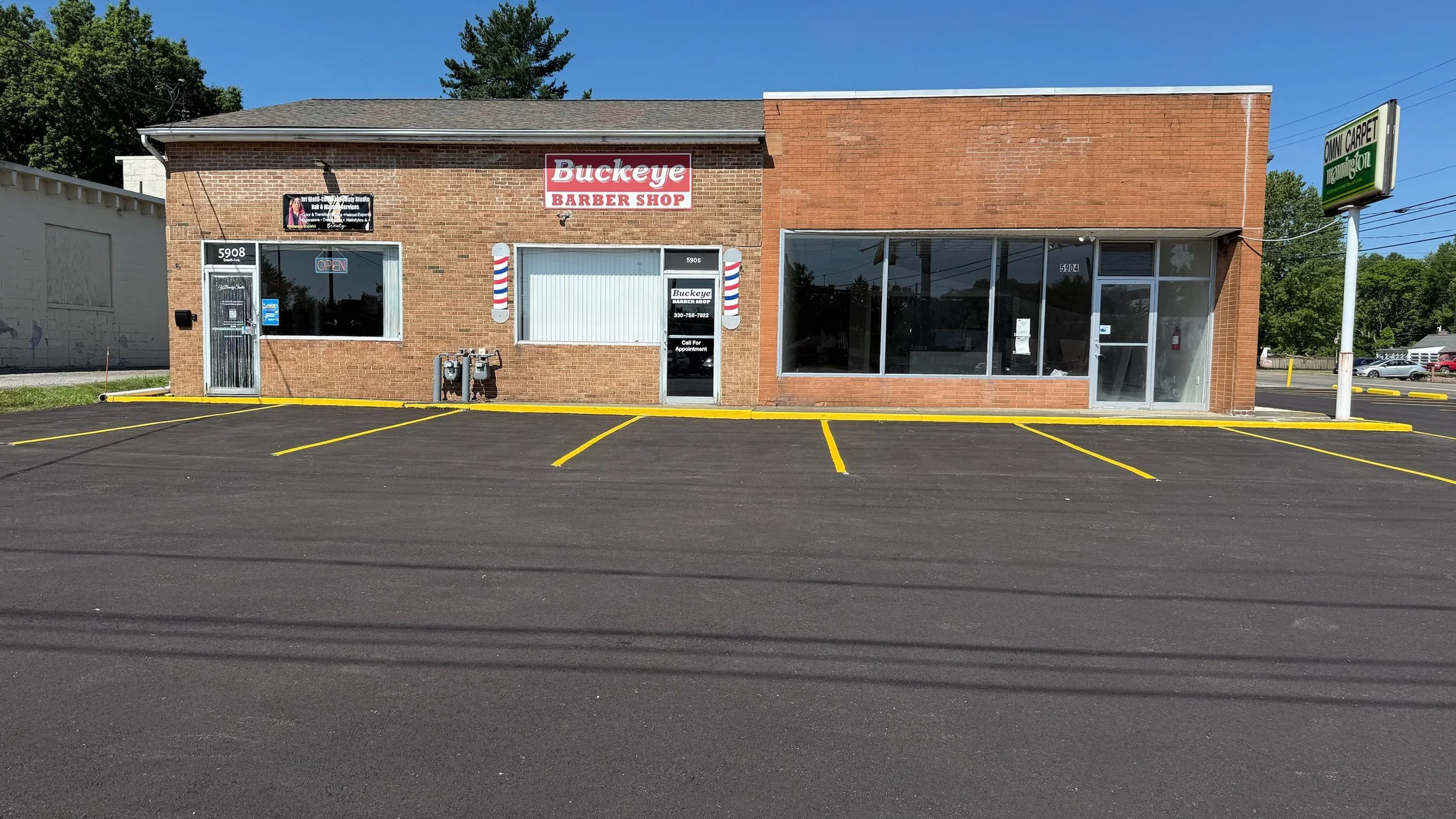Bricked building with signs for Buckeye Barber Shop and Omni Carpet Cleaning, a large front window, and empty parking spaces in front.