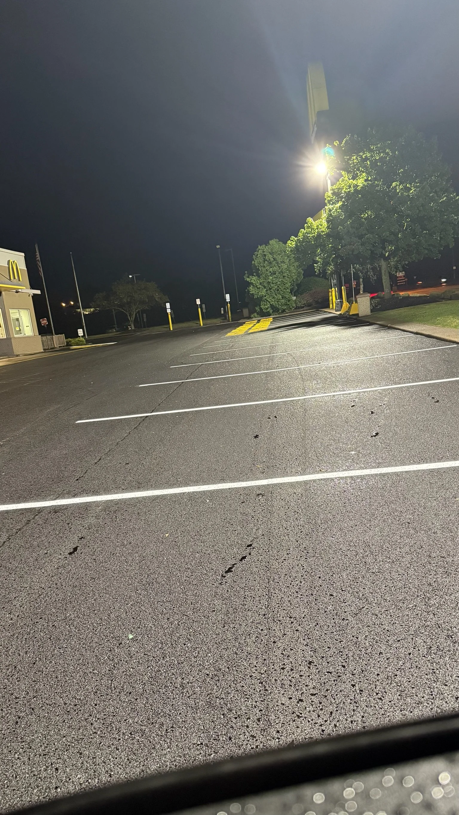 Empty parking lot at night outside a fast-food restaurant, illuminated by a bright streetlight, with some trees and a building in the background.