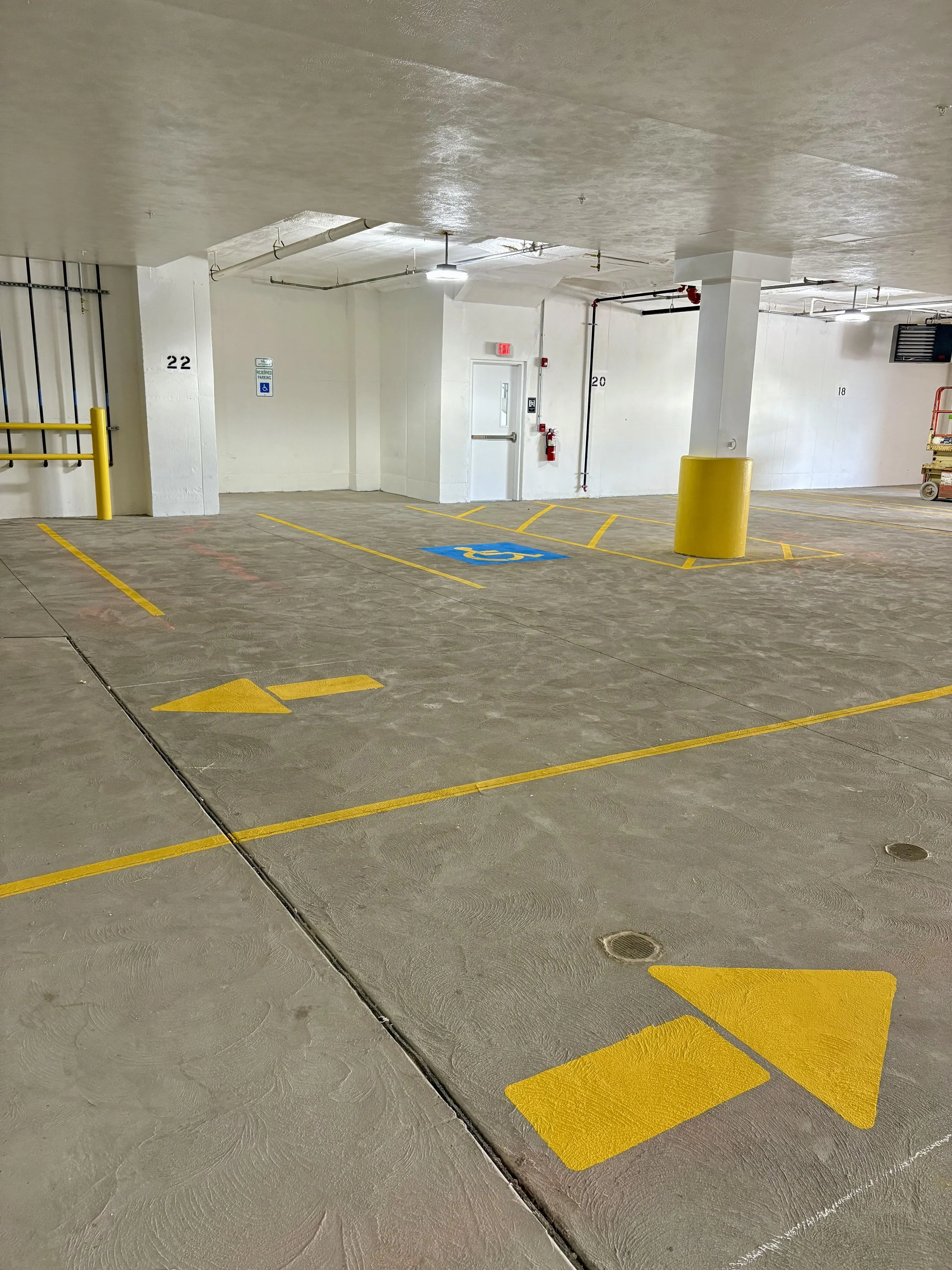 Underground parking lot with yellow parking space lines, a handicapped parking space marked blue, and parking space number 22 visible. There is a white door with an exit sign above it, a fire extinguisher, and a yellow safety barrier post.