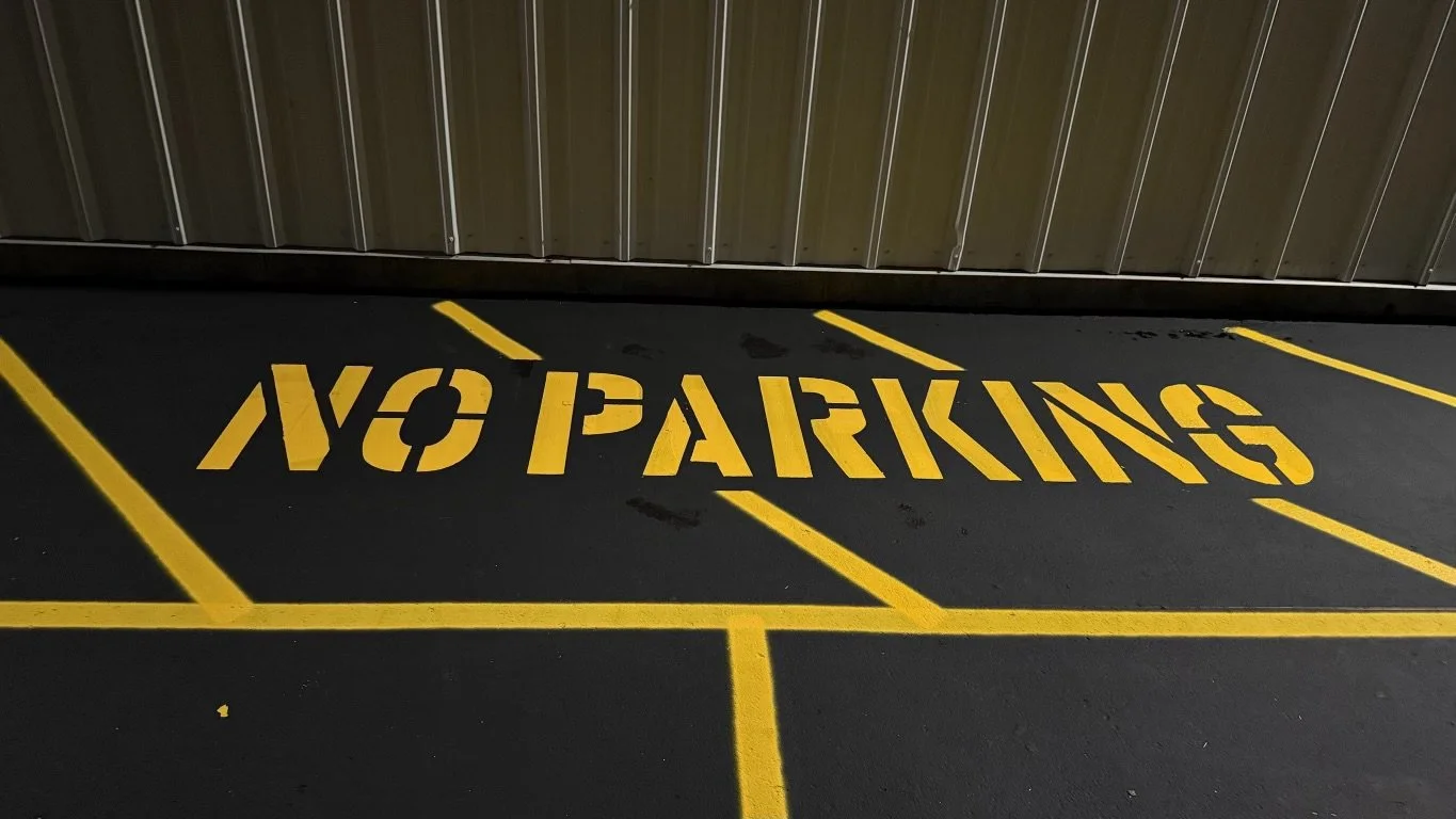 A parking lot with a yellow painted 'NO PARKING' sign on the ground. The sign is slightly tilted and partially obscured by yellow parking lines. There is a metal wall in the background.