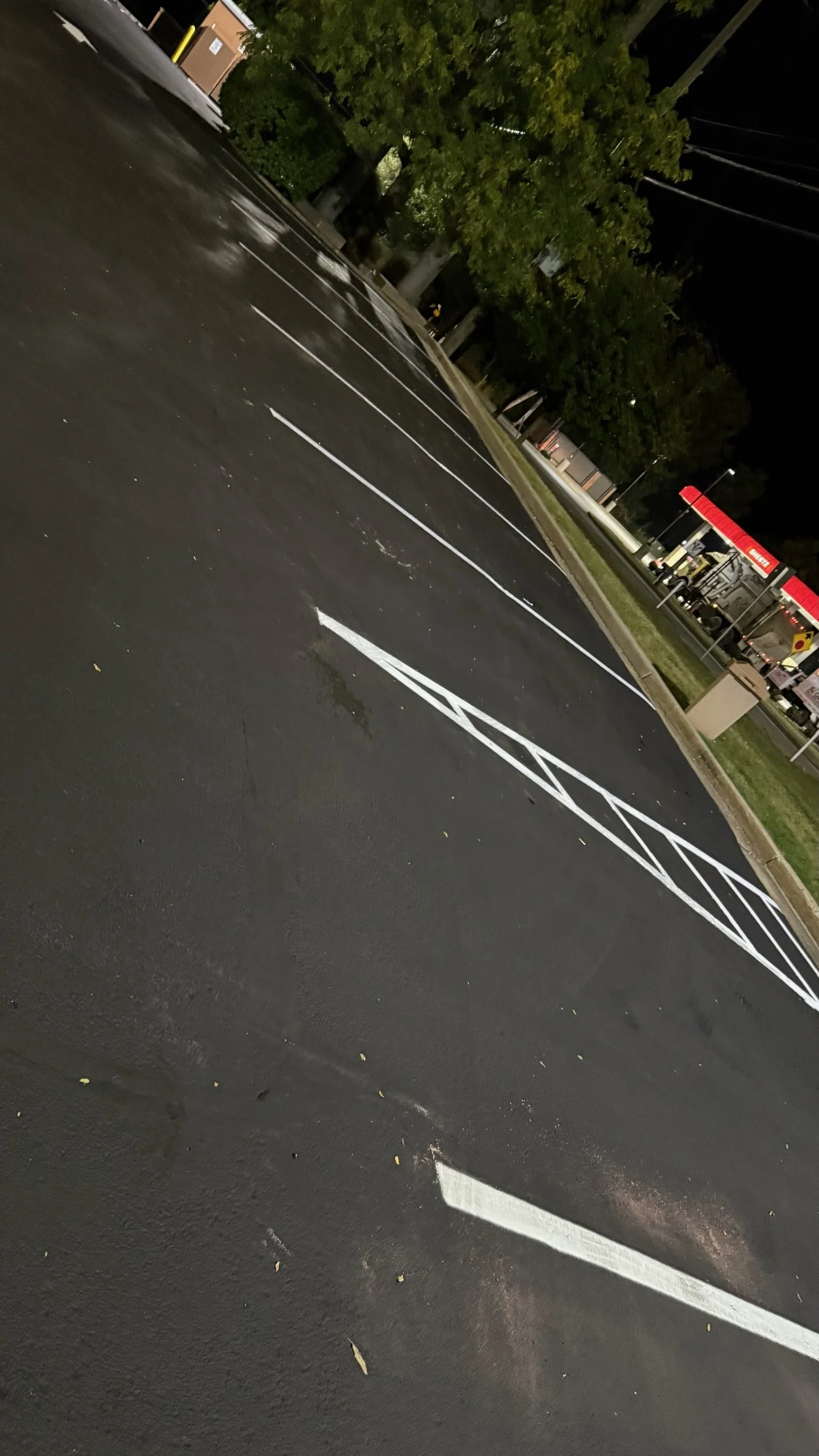 Empty parking lot at night with marked parking spaces, trees, a store with a red awning, and a sidewalk.