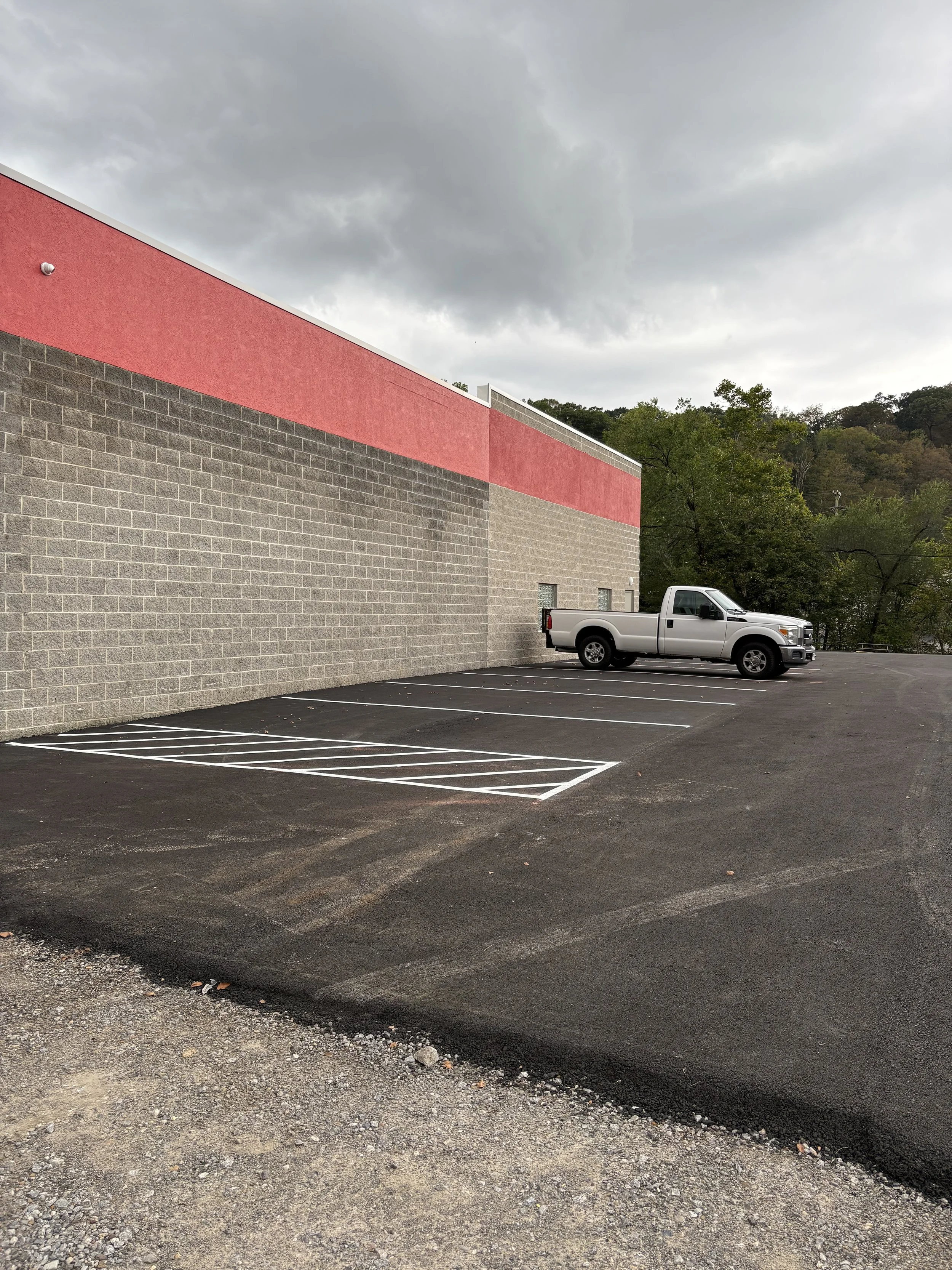 Empty parking lot with a white pickup truck parked near a building with a red upper wall and gray brick lower wall, surrounded by trees and cloudy sky.