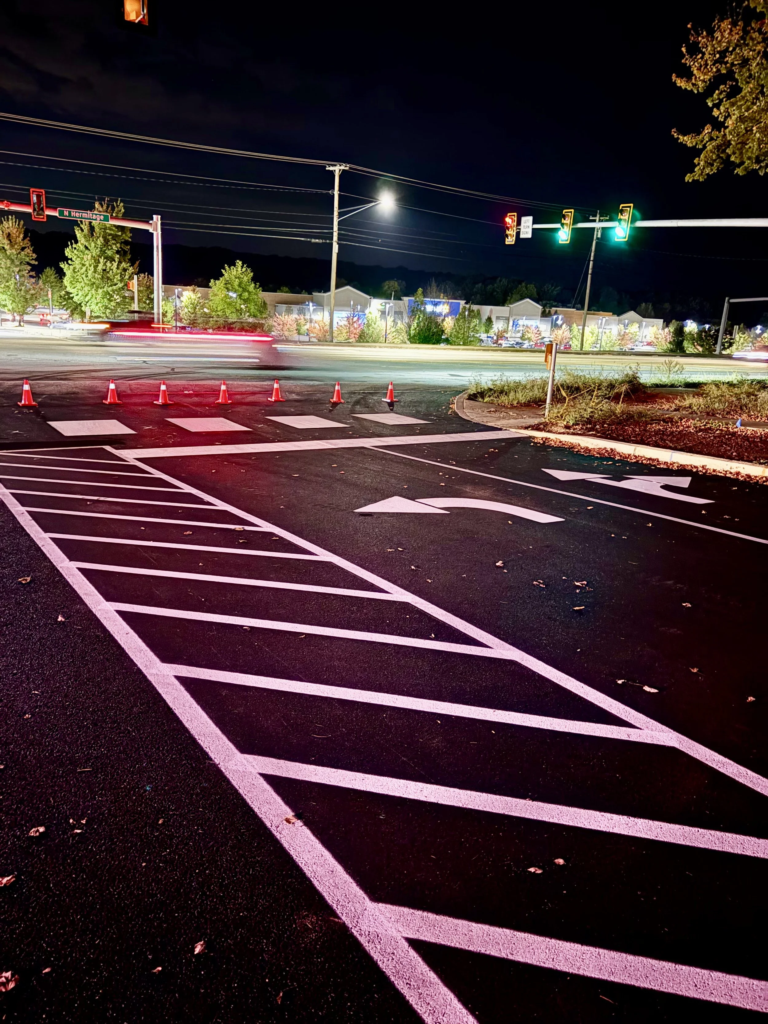 Nighttime view of a street with traffic lights, some cars moving, and a crosswalk. There are orange cones blocking part of the road, and a designated bicycle lane with pink markings in the foreground. Trees and buildings are visible in the background
