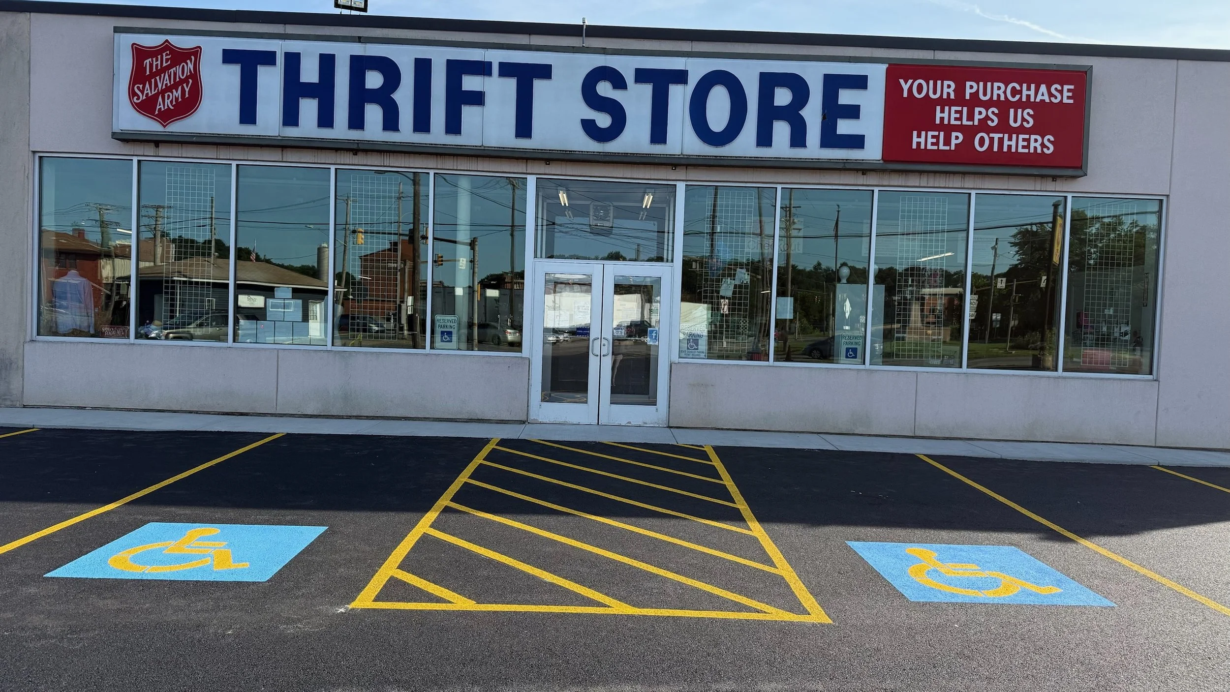 Front view of a thrift store with a sign reading 'Thrift Store' in large blue letters, a Salvation Army logo, and a red sign stating 'Your purchase helps us help others.' There are parking spaces in front, including designated accessible parking spac