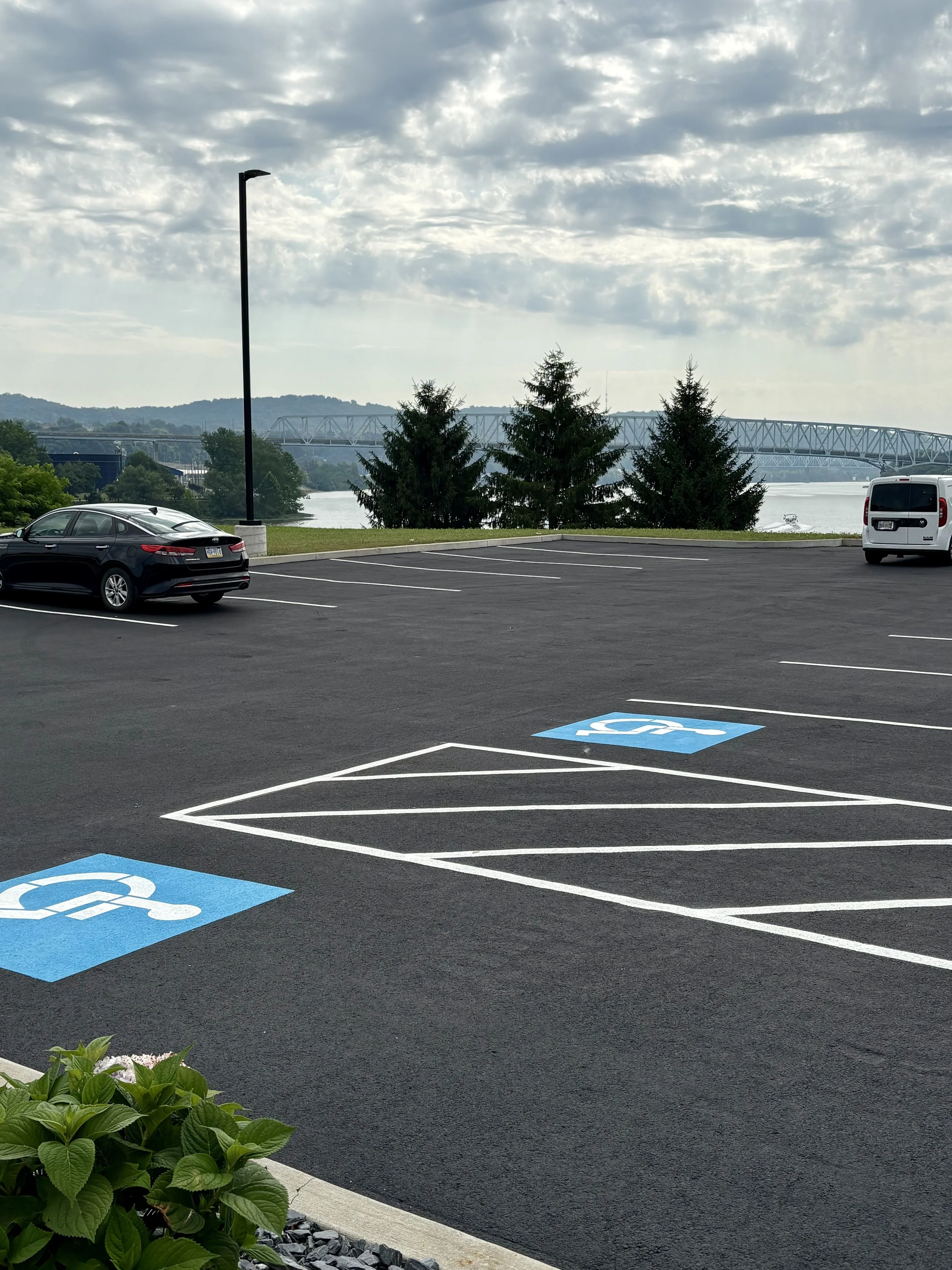 Empty parking lot with designated handicap accessible parking spots marked in blue and white, near a waterfront with trees, a bridge, and cloudy sky in the background.