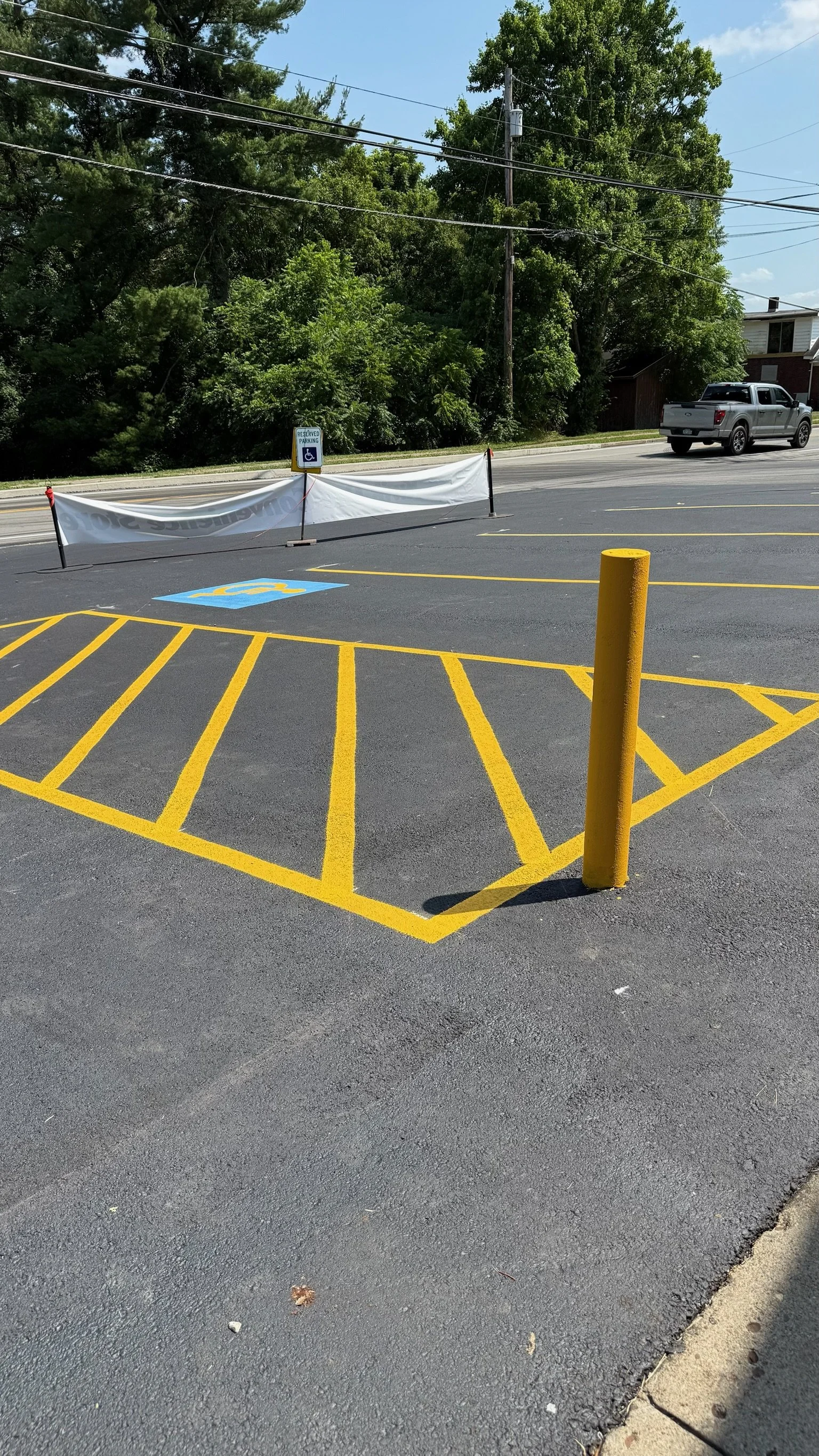 Empty parking lot with yellow painted lines, a handicapped parking spot, a yellow pole, and a barrier with a sign indicating reserved parking for handicapped. In the background, trees, power lines, and a parked vehicle are visible.