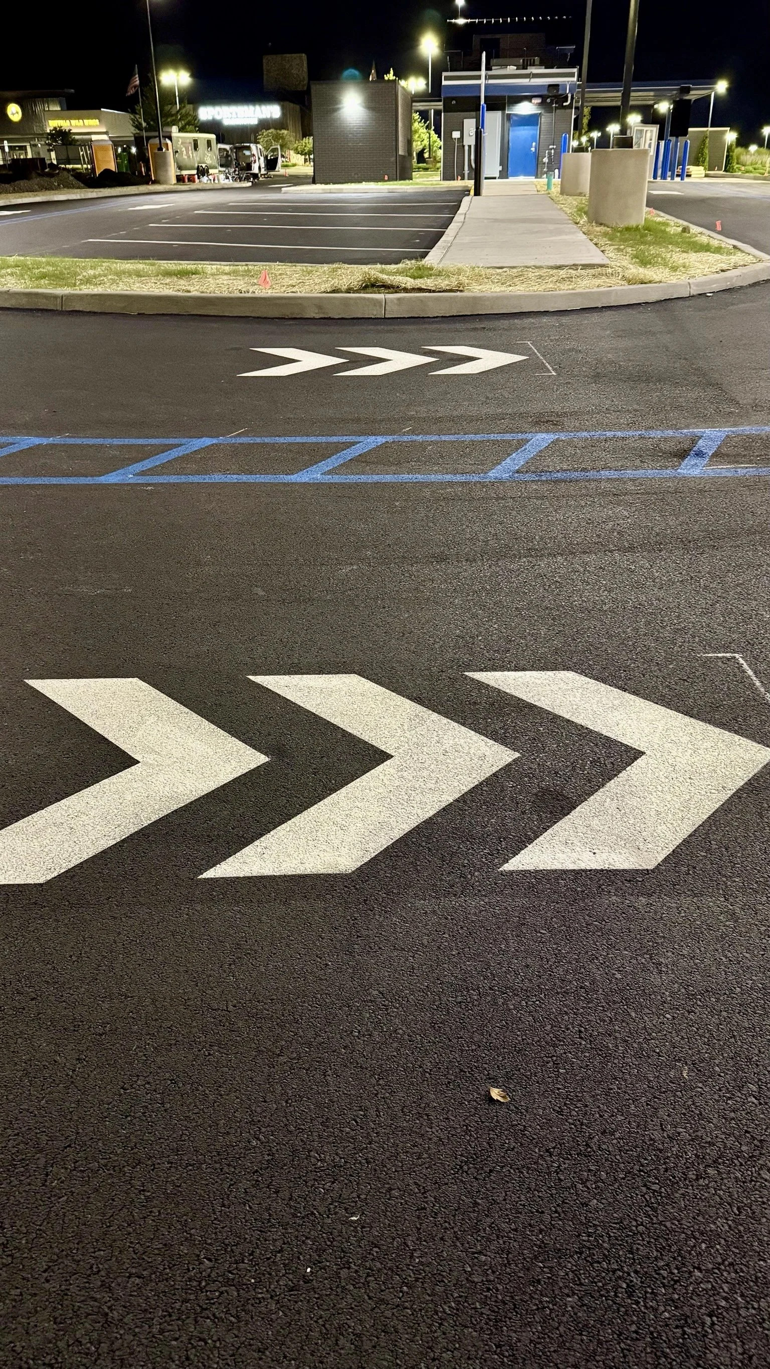 Nighttime view of a parking lot with white directional arrows painted on the asphalt, a curb, grass, and a sidewalk leading to small buildings, with lighting and some vehicles in the background.
