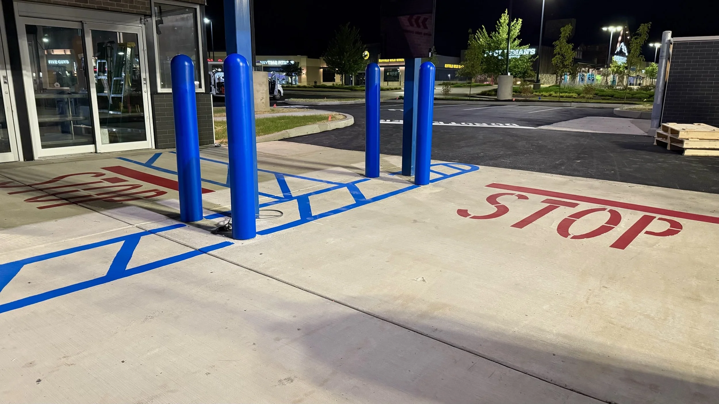 Empty parking lot with a designated handicapped parking space marked with blue lines and four blue metal posts, with a red 'STOP' painted on the concrete sidewalk outside a commercial building at night.