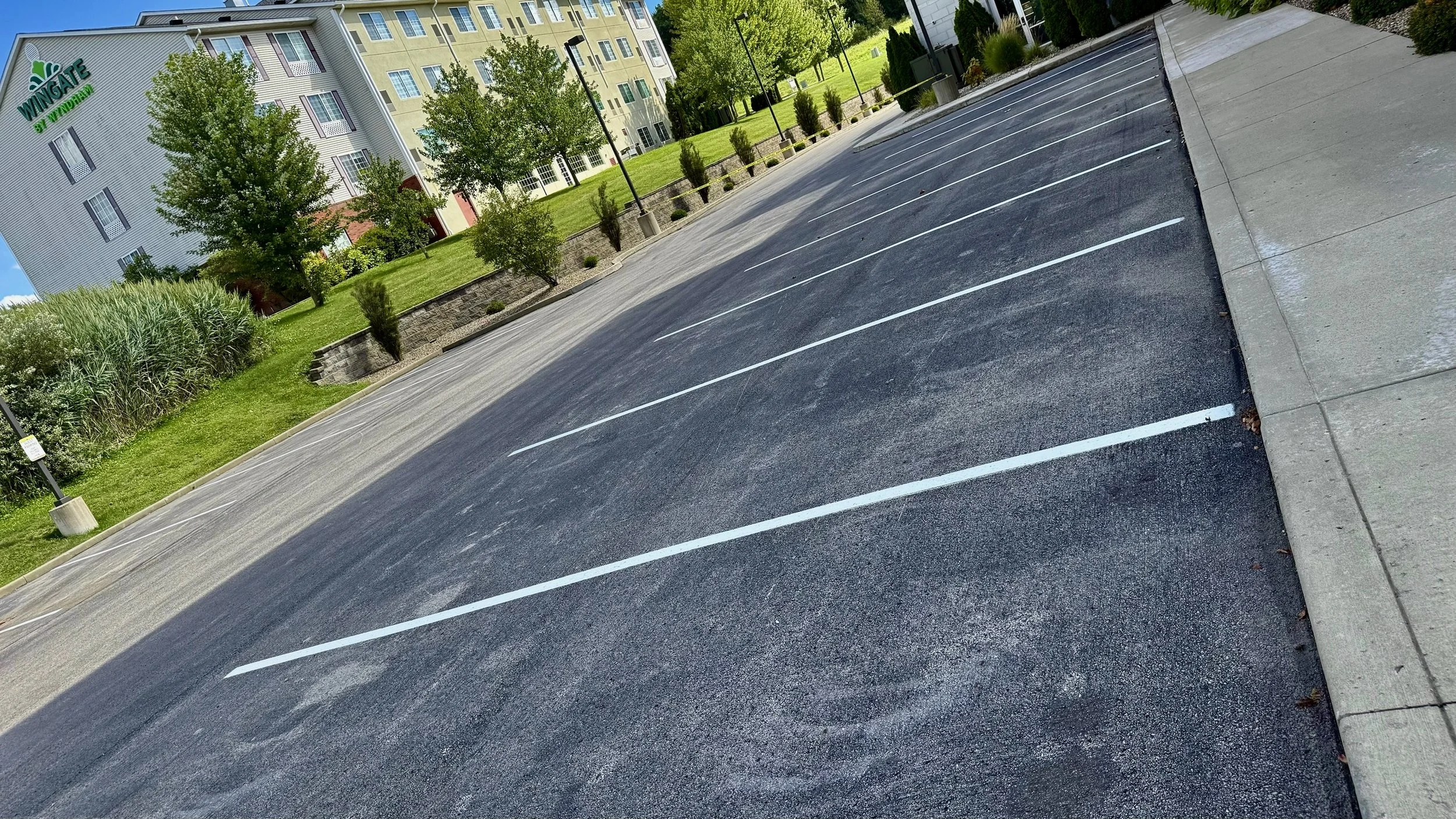 Empty parking lot with white painted parking spaces, sidewalk on the right, green grass, trees, and residential buildings in the background.