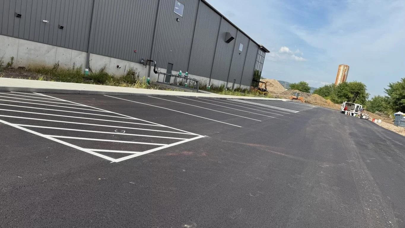 Empty parking lot with freshly paved asphalt and newly painted parking lines next to an industrial building, under a partly cloudy sky.