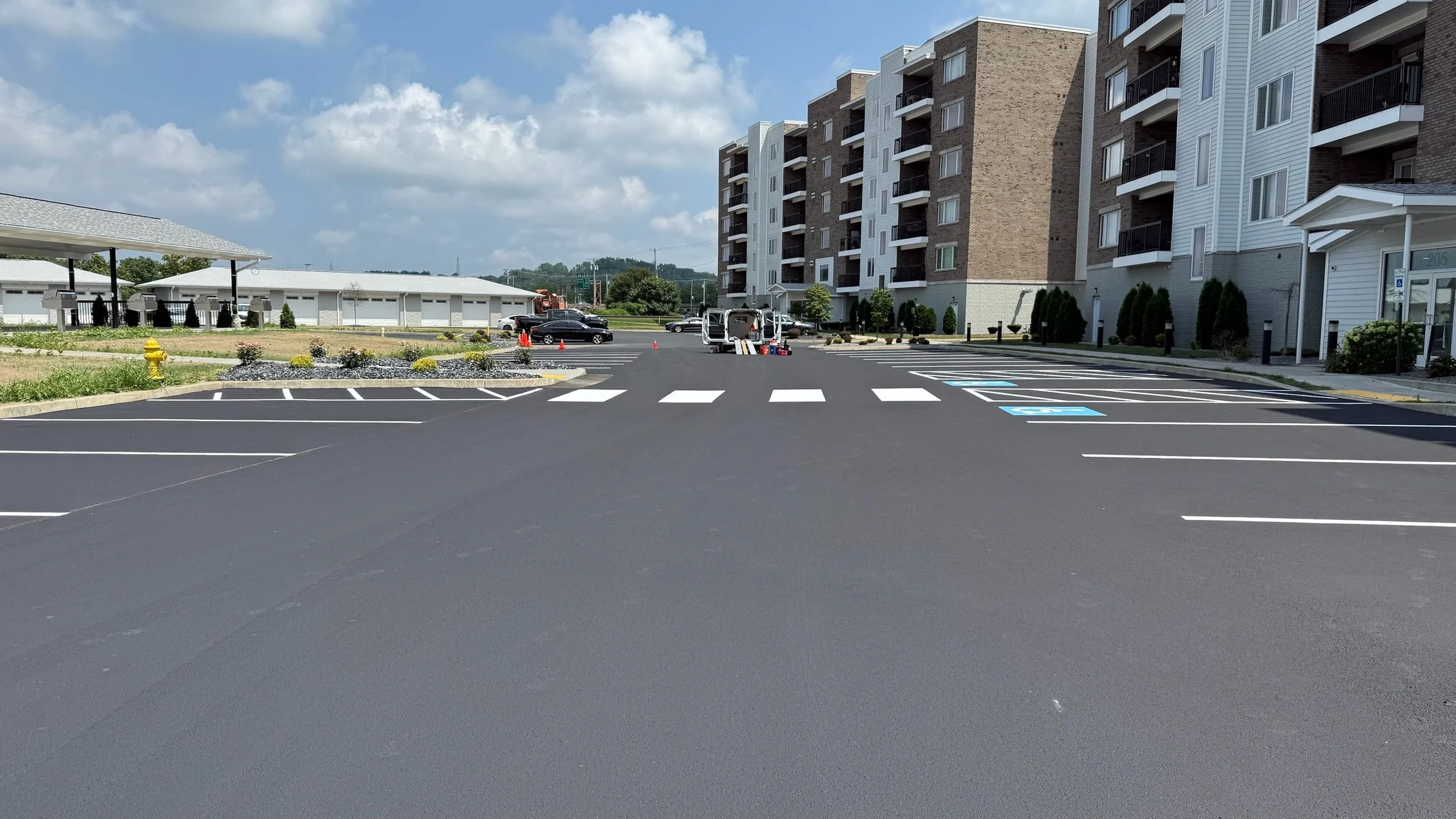 Empty parking lot with a crosswalk, corner parking spaces, and a large residential building in the background under a partly cloudy sky.