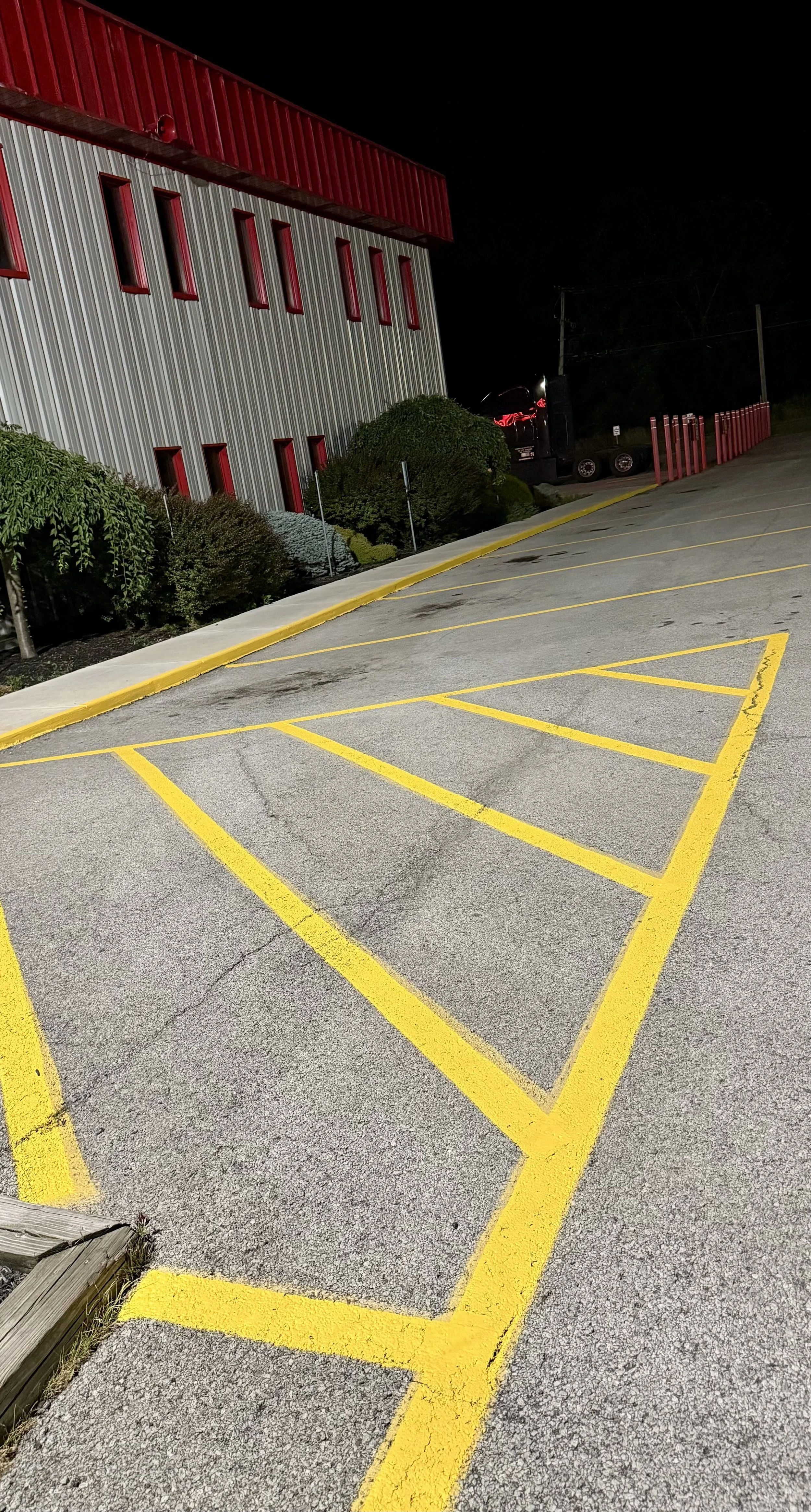 Empty parking lot with yellow striped parking spaces at night, adjacent to a red and gray building with small bushes along the side.
