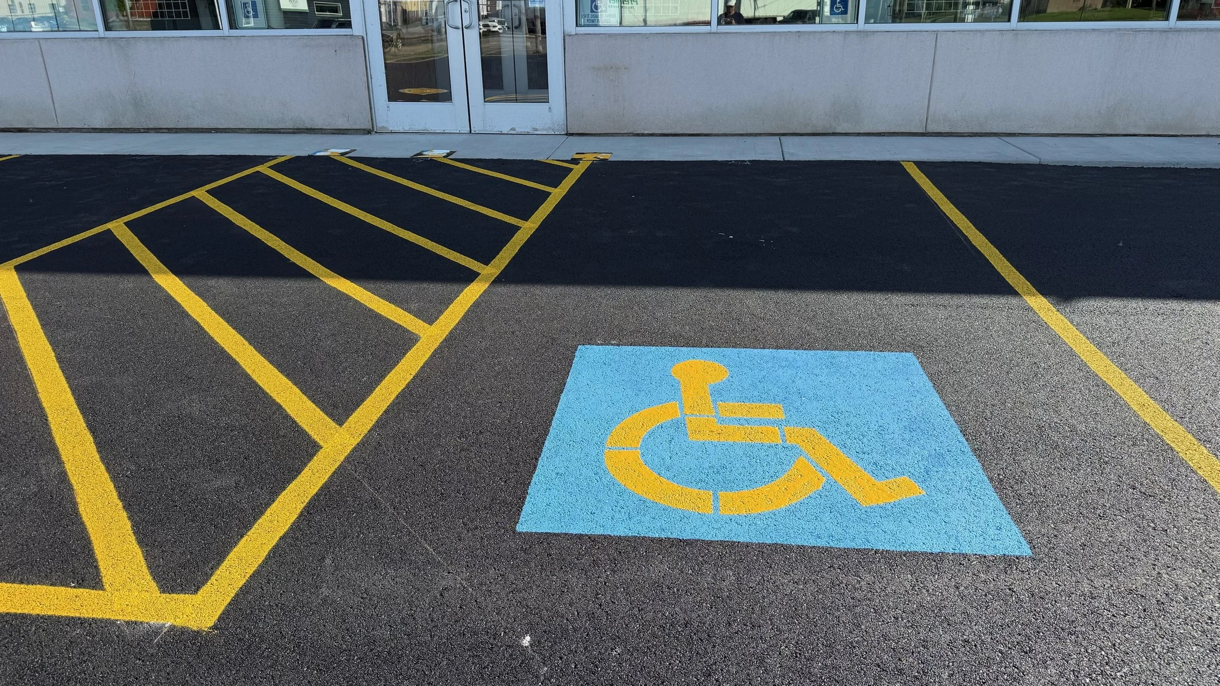 Designated accessible parking space with blue background and yellow wheelchair symbol, next to yellow striped lines indicating a no-parking zone in front of a building entrance.