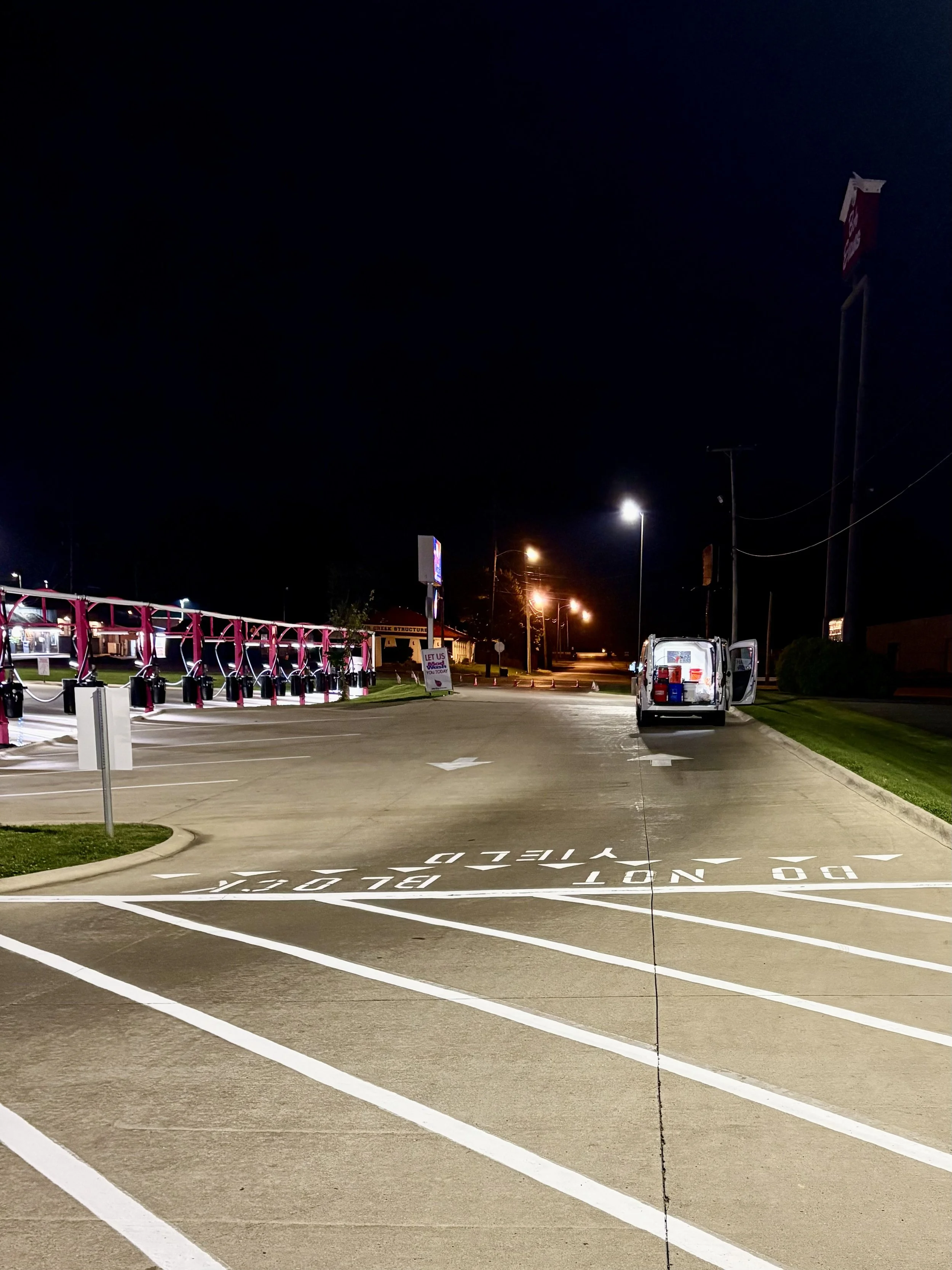 A parking lot at night with painted directional arrows and stopping points, a white van with open doors, and a row of gas pumps illuminated by exterior lights.