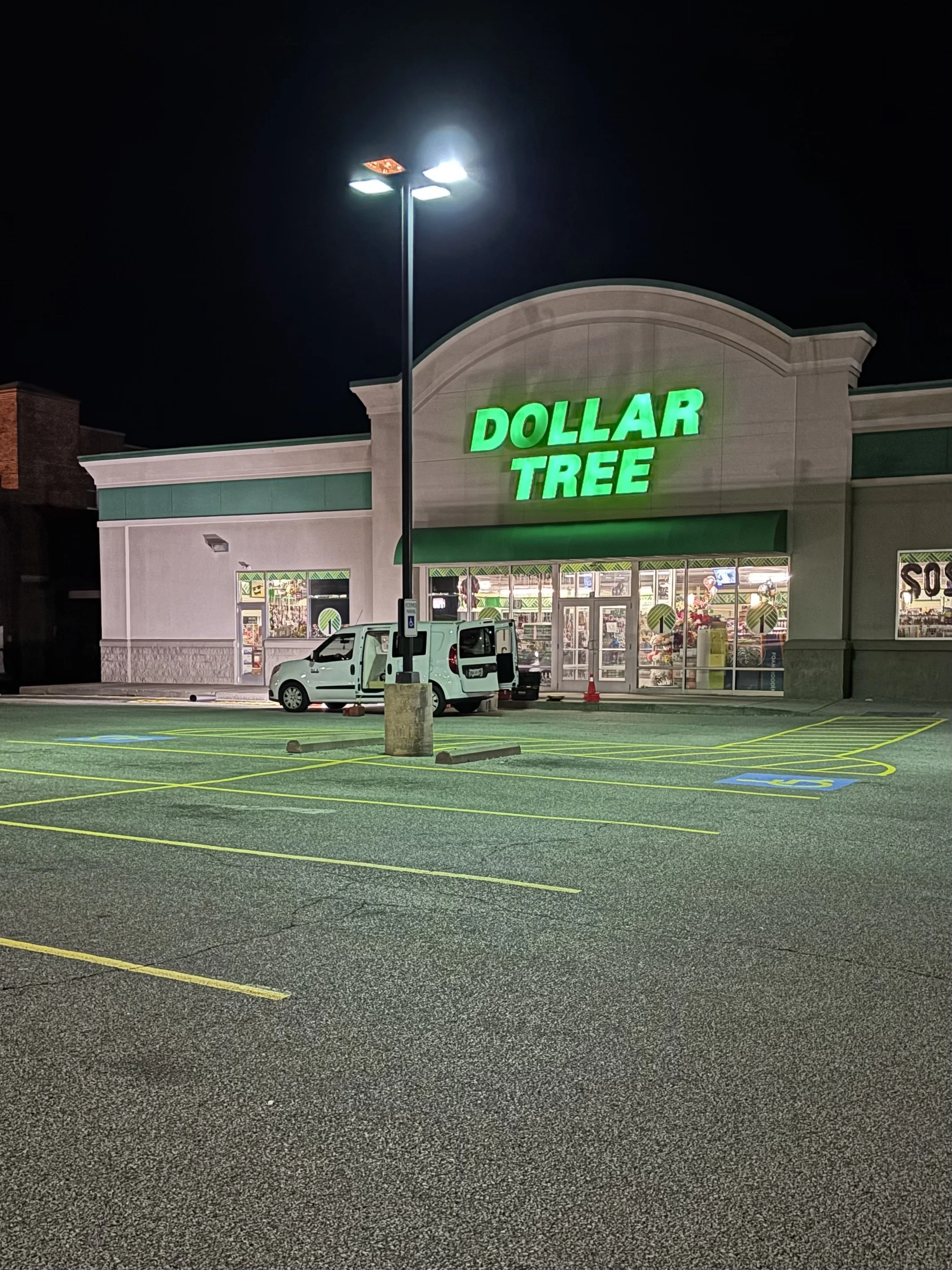 Nighttime view of a Dollar Tree store with empty parking lot, a white van parked in front, and a tall light pole illuminating the area.