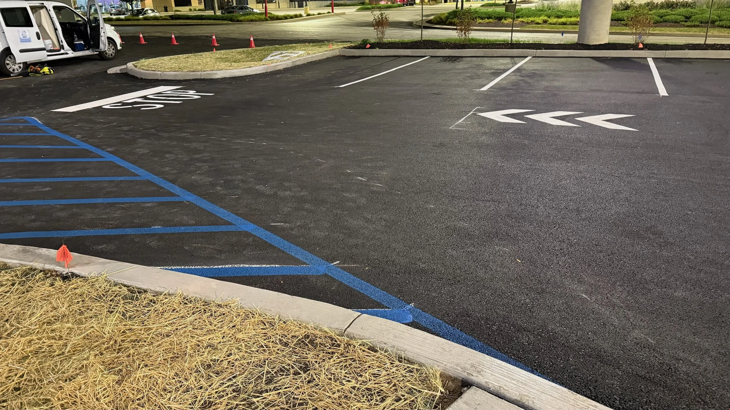 Empty parking lot with freshly painted blue and white parking lines, including a disabled parking space, and a small grassy area with mulch and a red flag marker. There are orange cones in the distance and a white van parked nearby.
