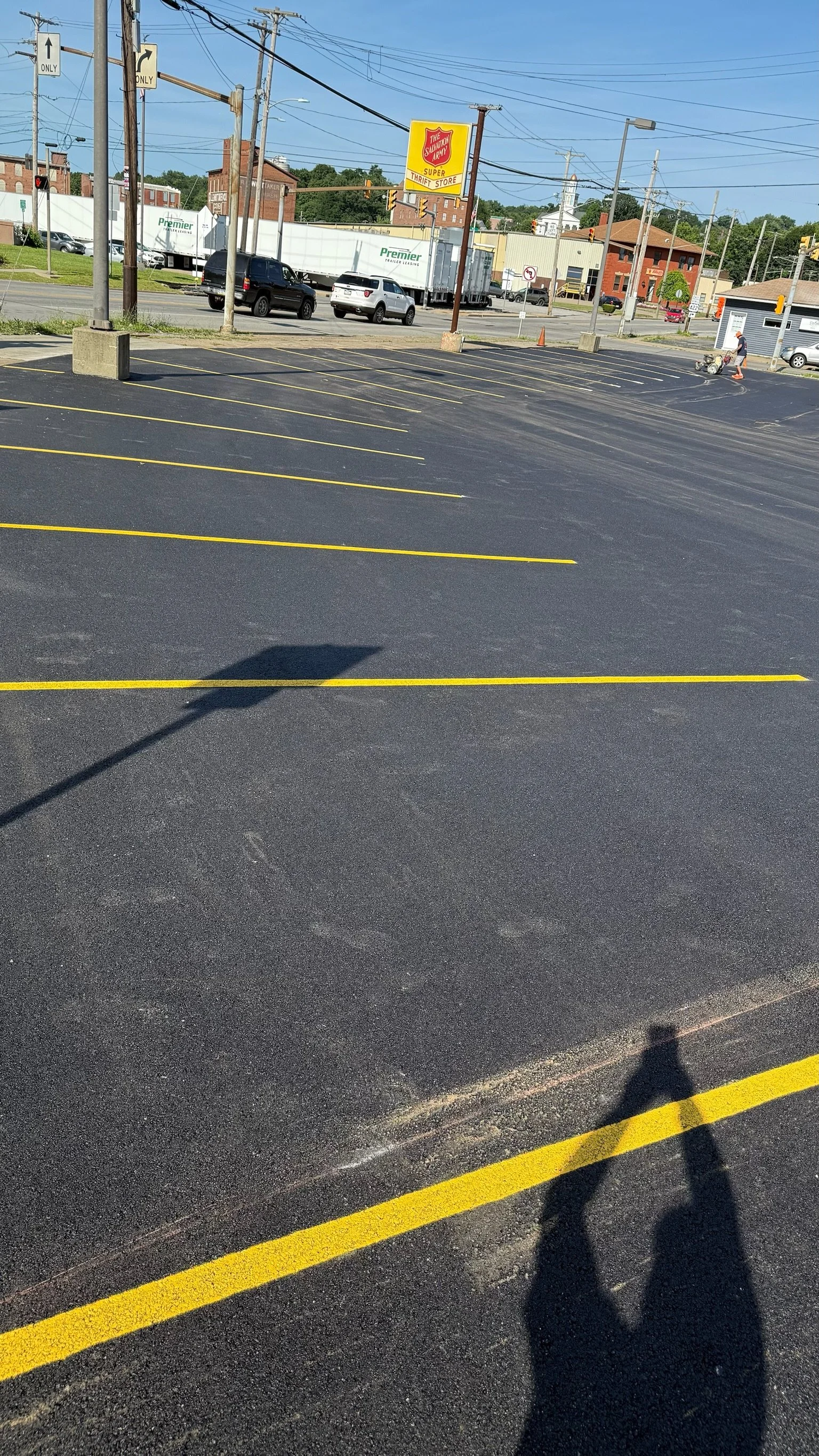 Empty parking lot with freshly painted yellow parking lines, a person pushing a shopping cart, a sign for The Salvation Army thrift store, and cars driving on the street with utility poles and buildings in the background.