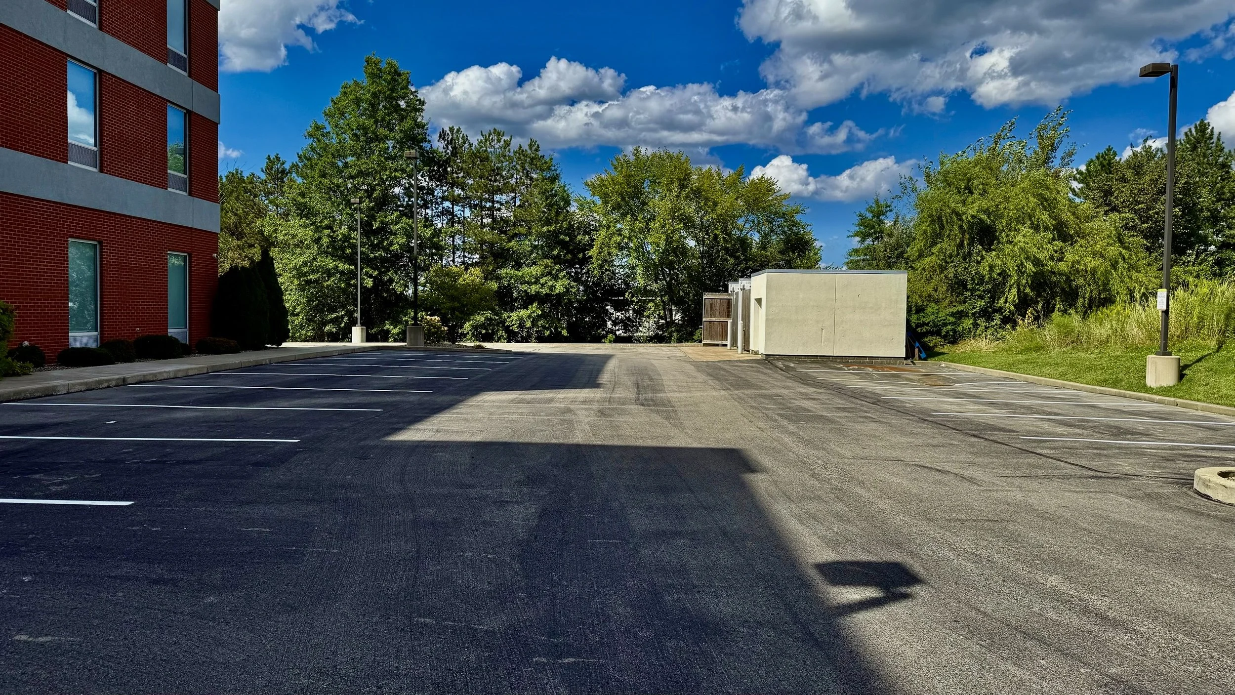 Empty parking lot next to a red brick building, with trees and greenery in the background under a partly cloudy blue sky.